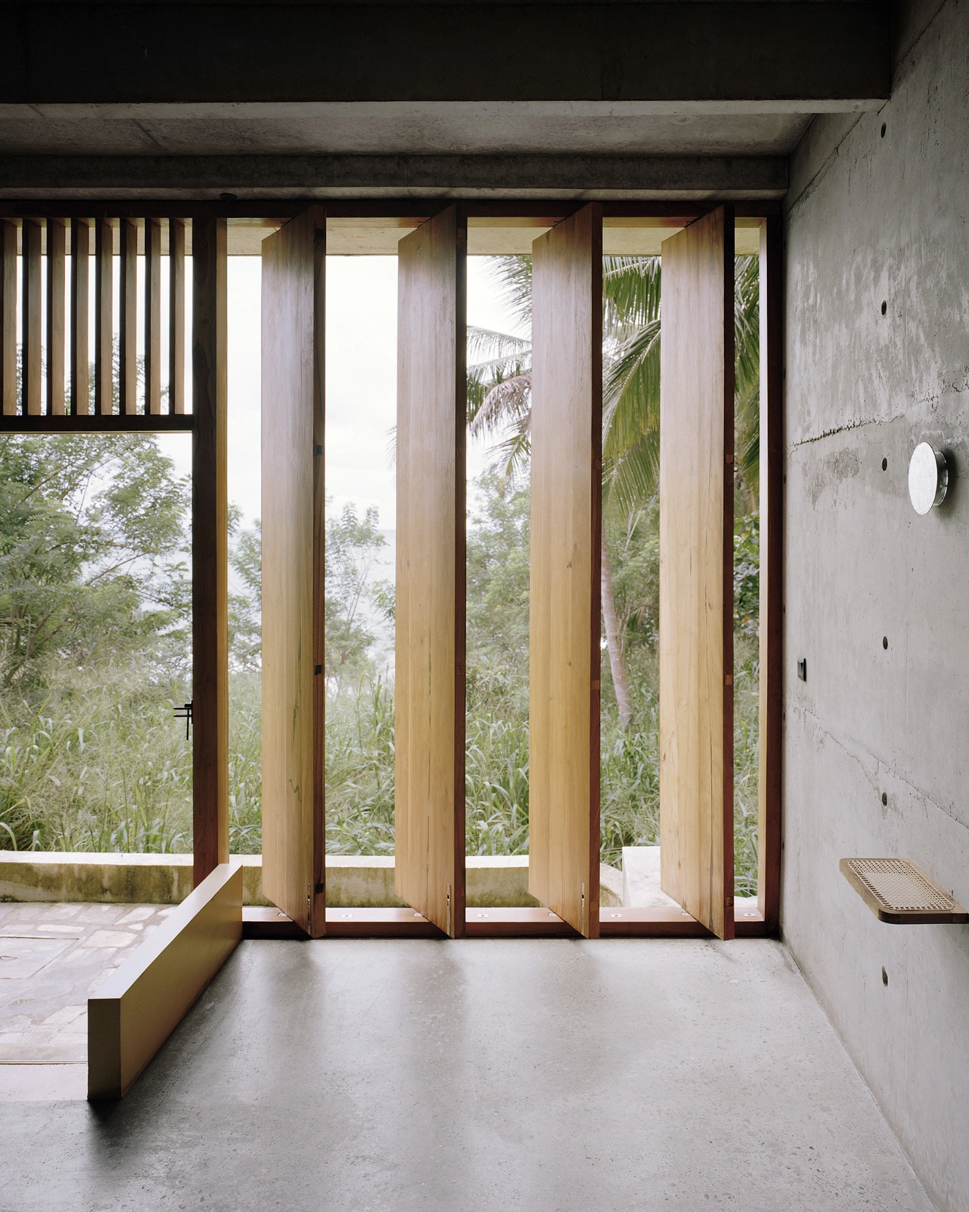 057 Guesthouse Hiriketiya by Atelier Abraha Achermann — A view through a series of timber-framed glass doors opening onto a tropical landscape.