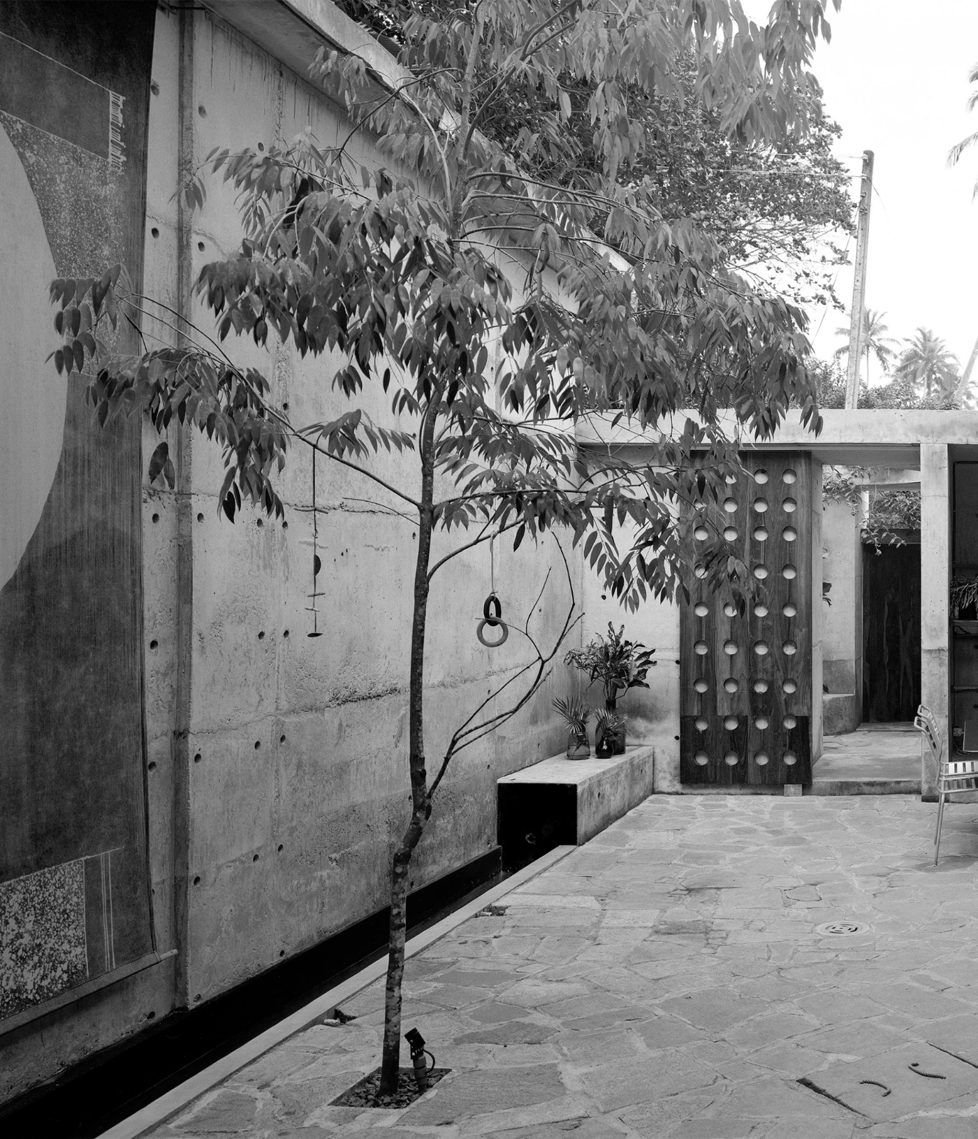 057 Guesthouse Hiriketiya by Atelier Abraha Achermann — A monolithic concrete courtyard wall dominates the left frame, its surface marked by horizontal board-formed striations and visible cylindrical bolt holes...