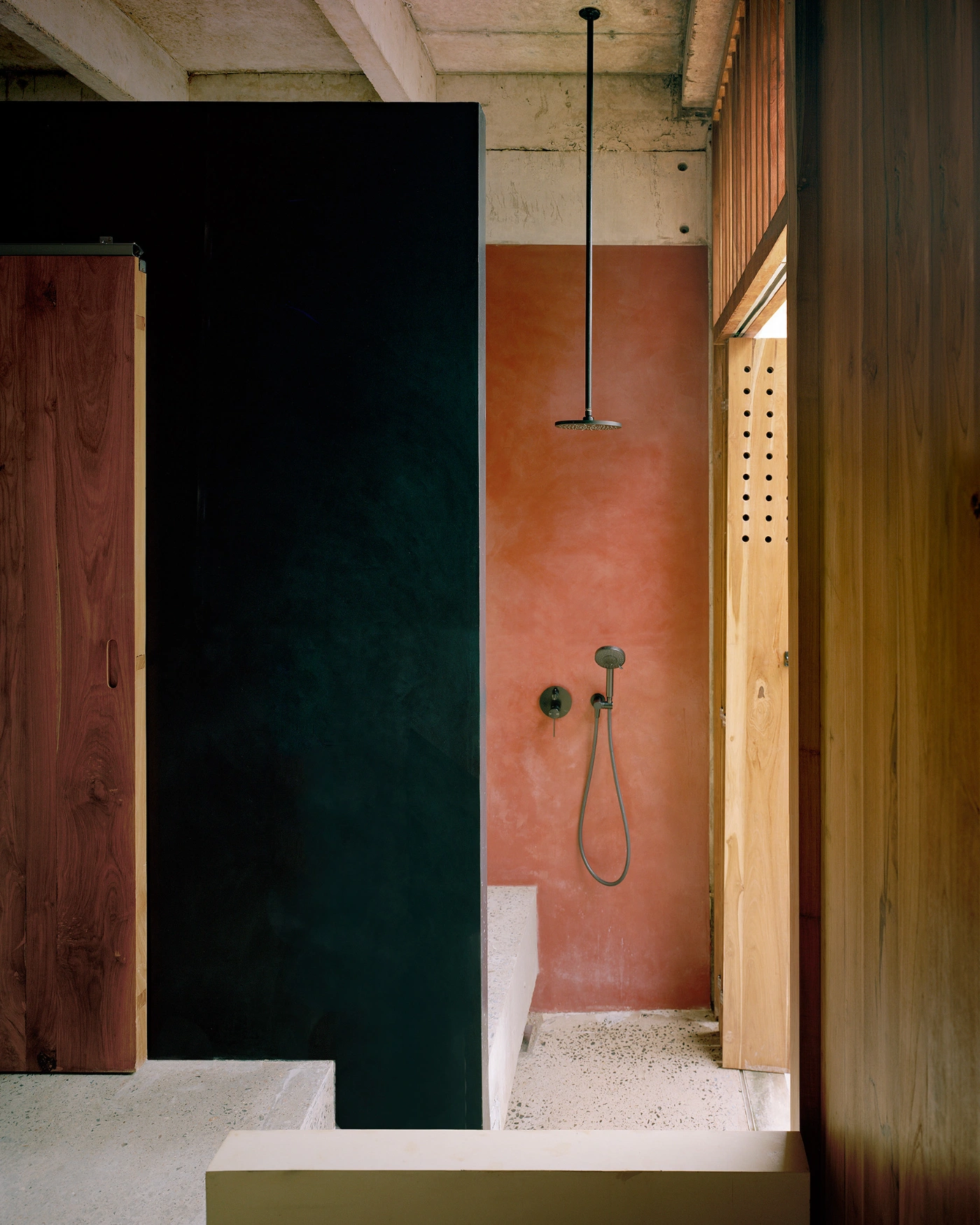 057 Guesthouse Hiriketiya by Atelier Abraha Achermann — A narrow shower enclosure framed by colorful rendered walls in terracotta and deep teal, separated by a black board-marked concrete panel.