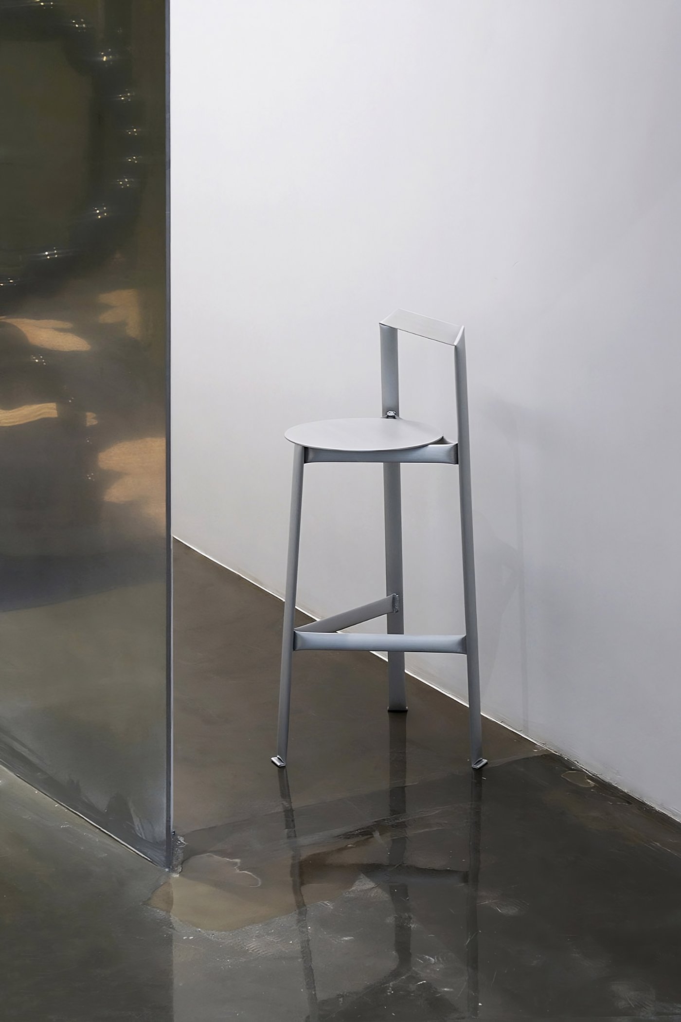 MONOCHROME by Ximi Li — A tall aluminum bar stool occupies the center of a polished dark floor, positioned against a white wall.
