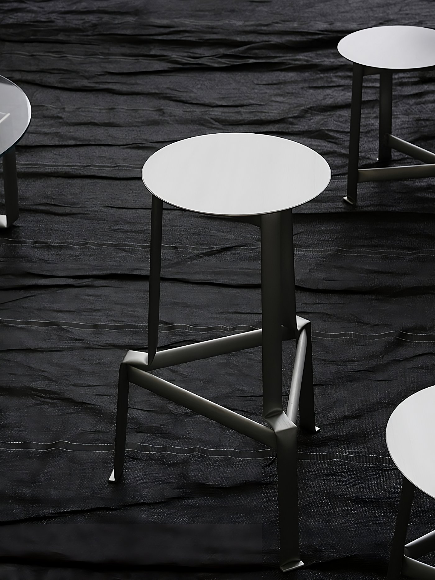 MONOCHROME by Ximi Li — Circular aluminum side table with white top mounted on welded black metal frame, positioned on dark textured floor.