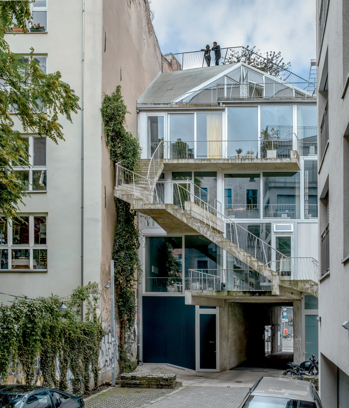 Brunnenstrasse 9 by Brandlhuber+ by Brandlhuber & Emde & Burlon — A diagonal concrete cantilever staircase with steel cable railings cuts across the narrow courtyard facade, creating a sculptural circulation element that...