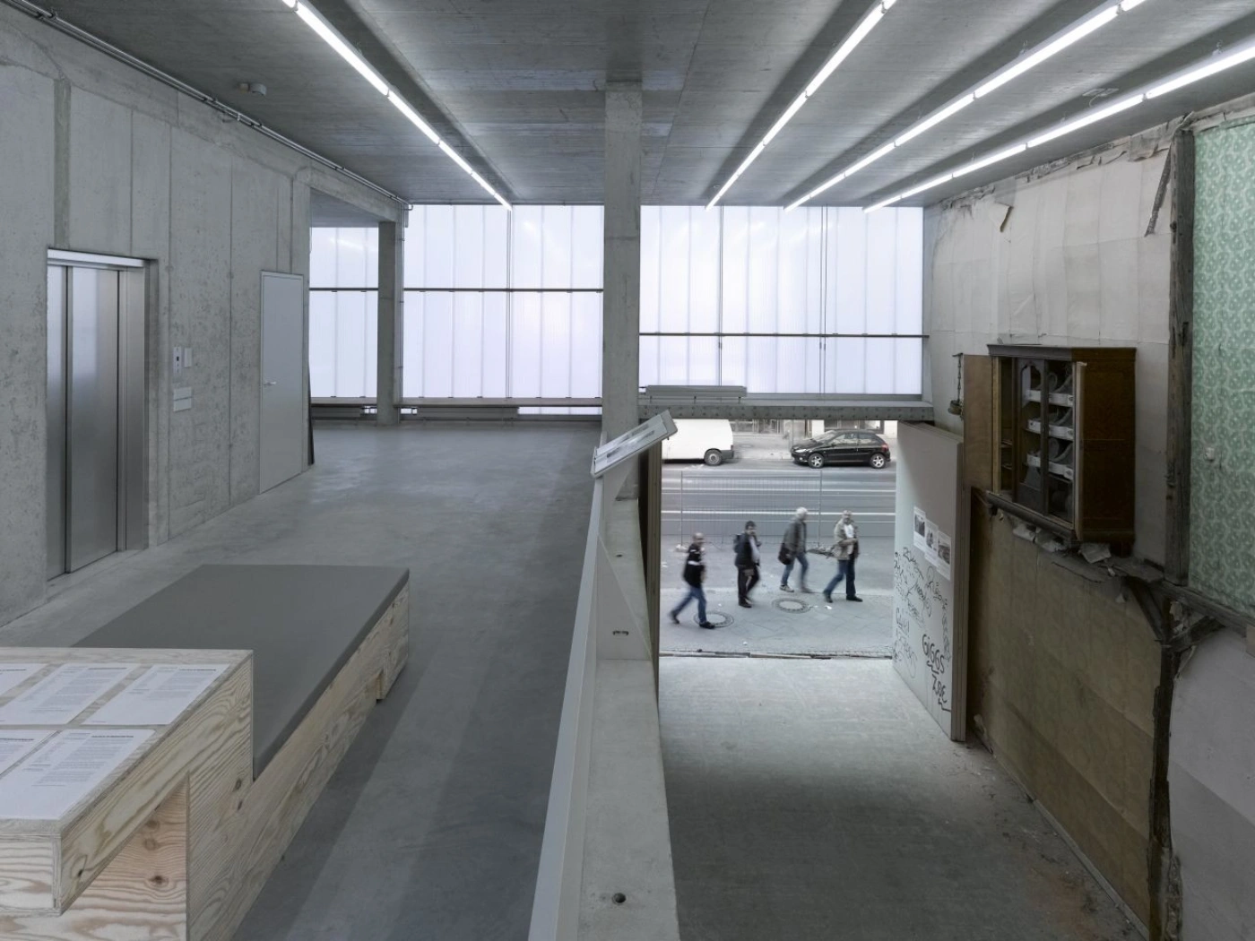 Brunnenstrasse 9 by Brandlhuber+ by Brandlhuber & Emde & Burlon — A passage-through threshold beneath upper floors frames a street-level view of parked vehicles and pedestrians.