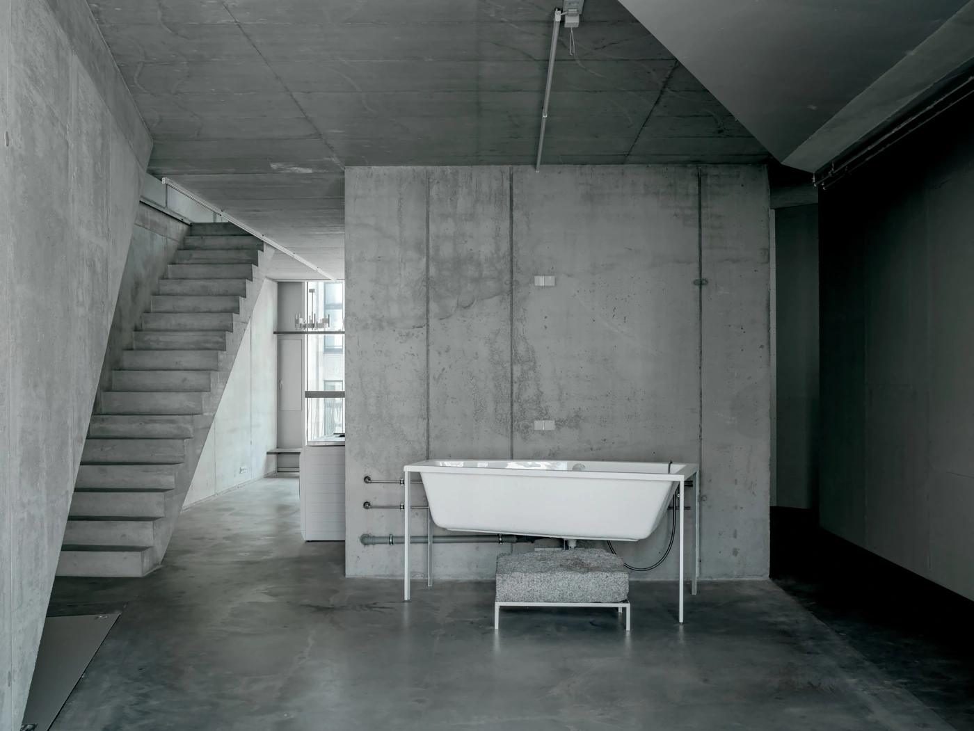 Brunnenstrasse 9 by Brandlhuber+ by Brandlhuber & Emde & Burlon — A bathroom installation within raw concrete interior space, featuring a white freestanding bathtub with exposed steel plumbing fixtures positioned centrally...