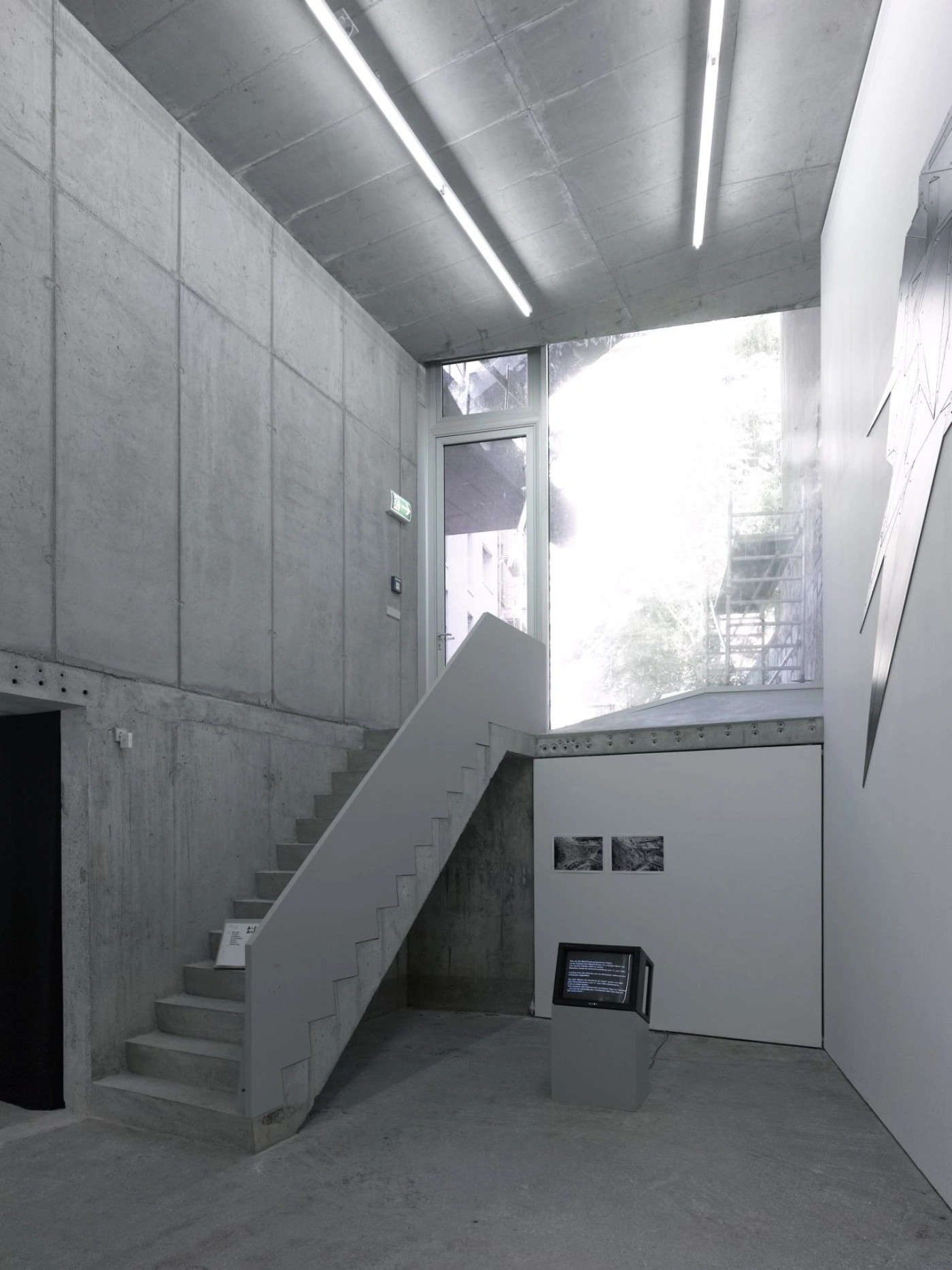 Brunnenstrasse 9 by Brandlhuber+ by Brandlhuber & Emde & Burlon — A concrete interior passage with a diagonal cantilever staircase ascending the left wall, its underside exposed to reveal structural depth.