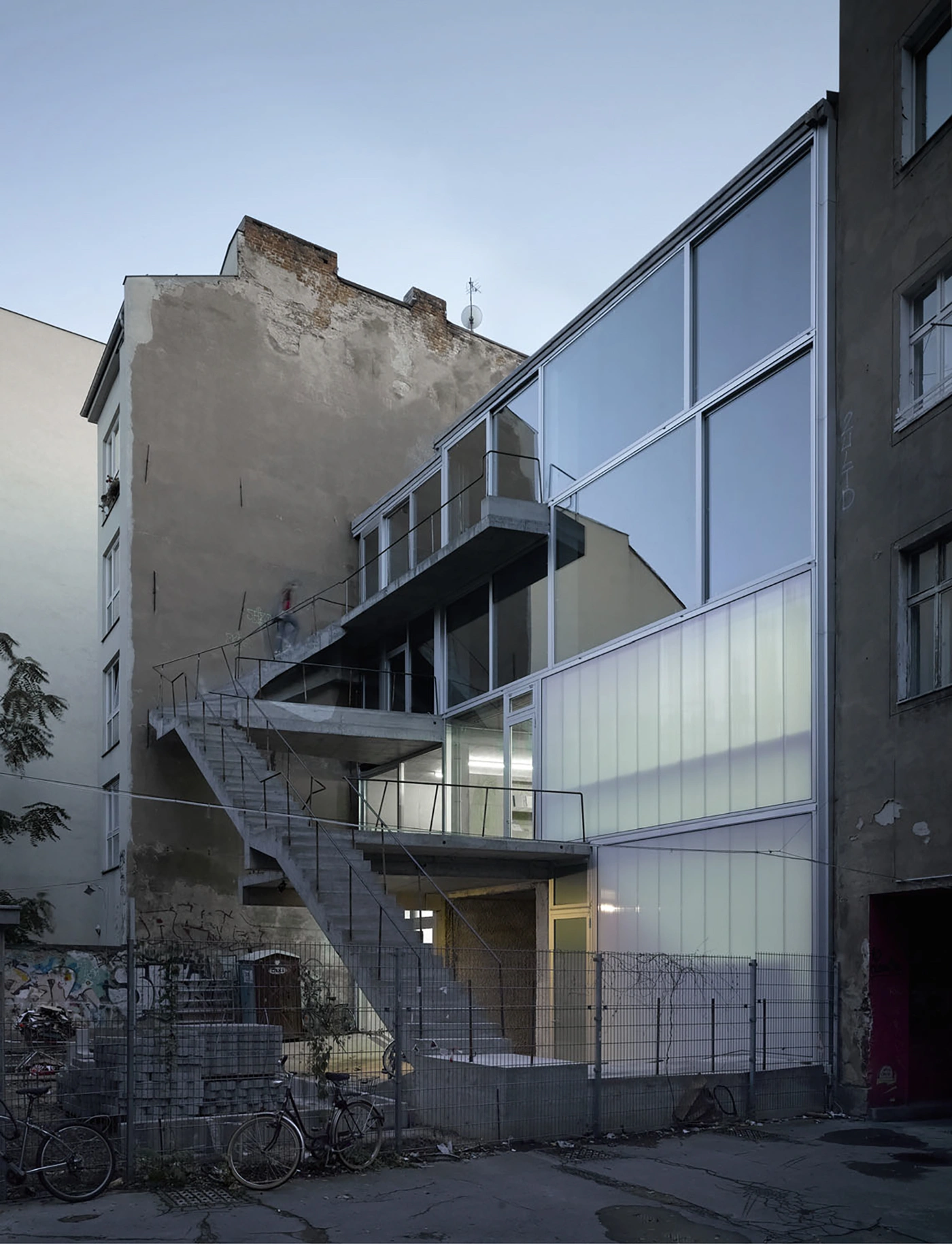 Brunnenstrasse 9 by Brandlhuber+ by Brandlhuber & Emde & Burlon — A diagonal concrete cantilever staircase with steel cable railings cuts across the weathered facade of a Berlin courtyard building, connecting multiple...