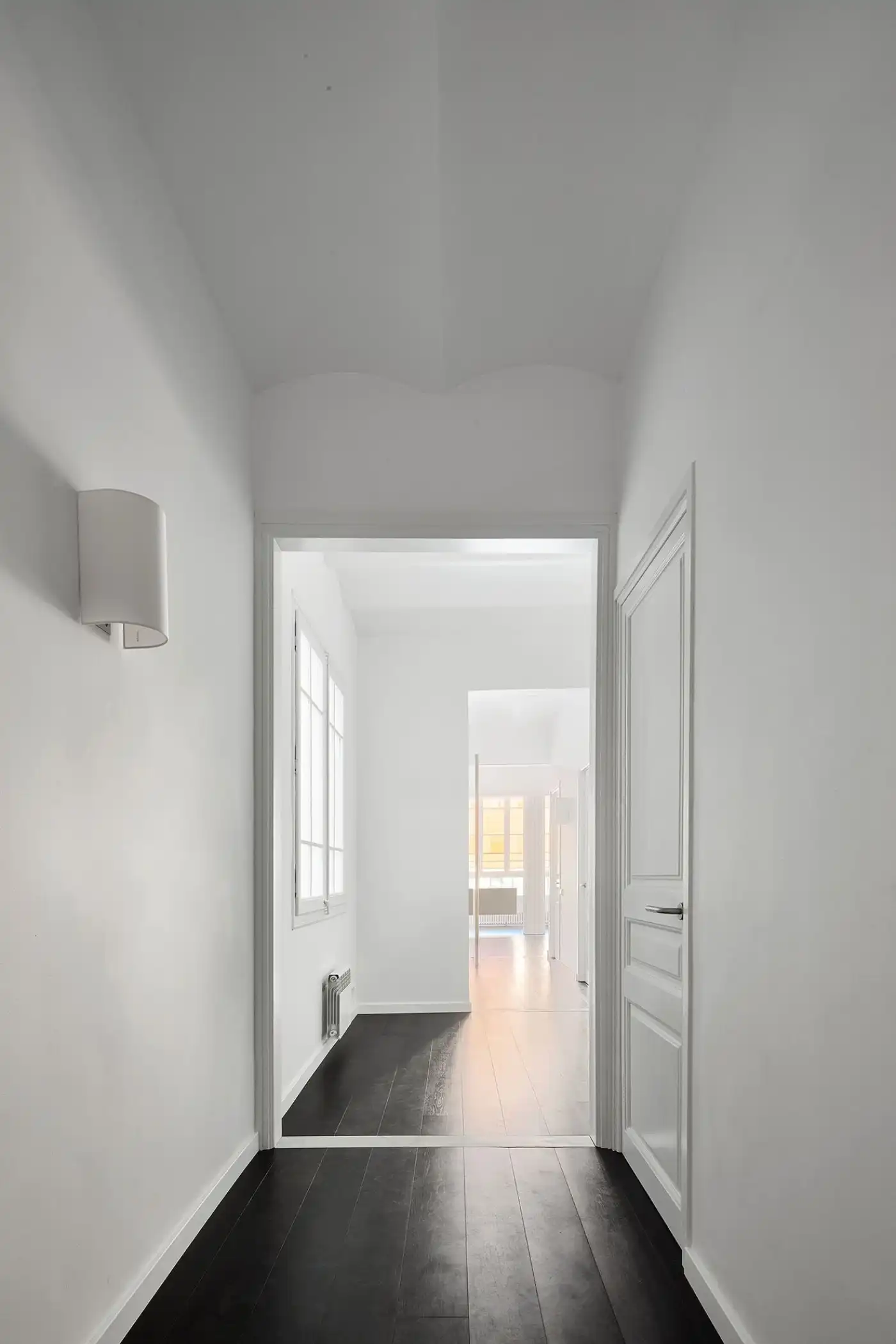 Flat in Eixample by Jaime Prous Architects — A rectilinear hallway extends through the Eixample flat, framed by white walls and a white ceiling with exposed concrete beam geometry.