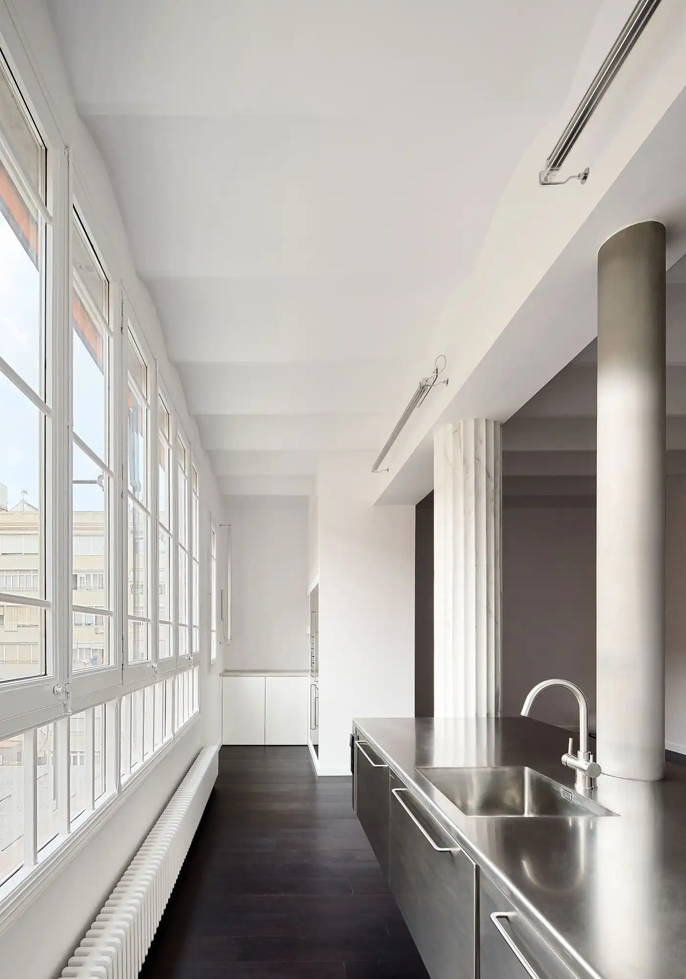 Flat in Eixample by Jaime Prous Architects — A galley kitchen extends along the western facade, where continuous multi-pane steel-framed windows frame a dense urban streetscape.