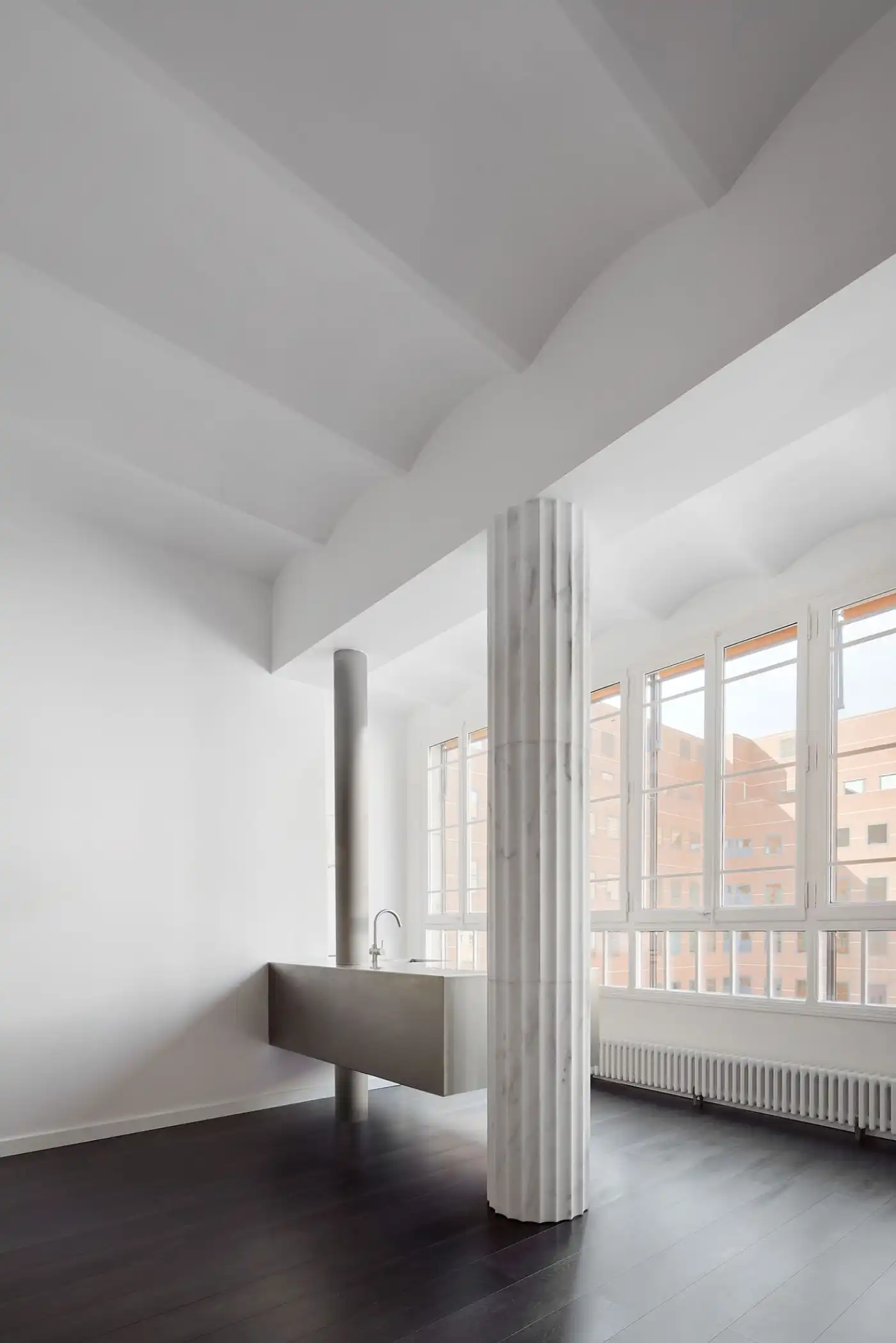 Flat in Eixample by Jaime Prous Architects — A cantilevered stainless-steel basin extends from a fluted cylindrical column that anchors the open-plan interior, positioning a functional element as...