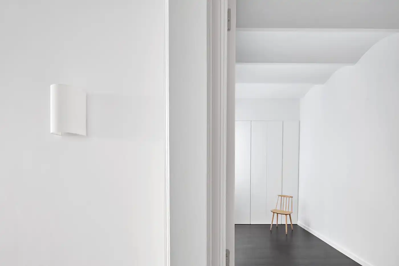 Flat in Eixample by Jaime Prous Architects — A minimalist corridor frames a wooden chair against white paneled walls and dark polished concrete flooring.