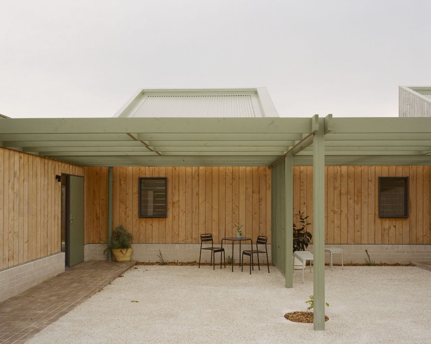 WPI Older Women’s Housing Project by Bright Studio by Studio Bright — Courtyard view of the shared housing project showing two timber-clad units separated by a sage-green steel pergola structure.