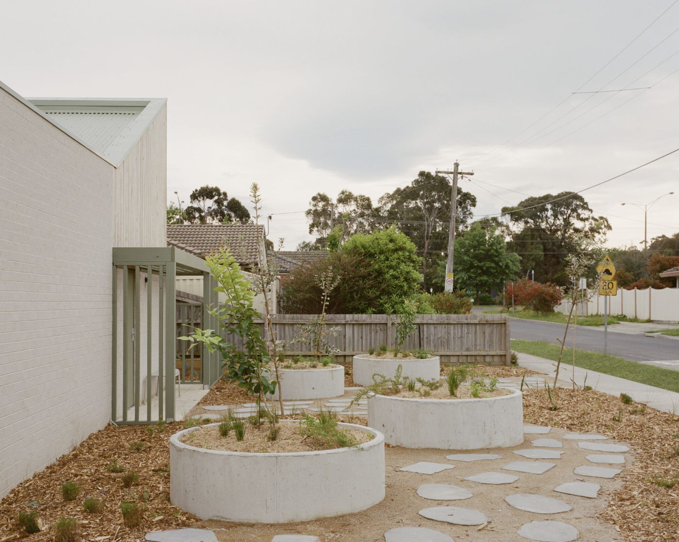 WPI Older Women’s Housing Project by Bright Studio by Studio Bright — The courtyard threshold features a progression of circular concrete planters with mixed plantings arranged across gravel and stepping-stone paving.