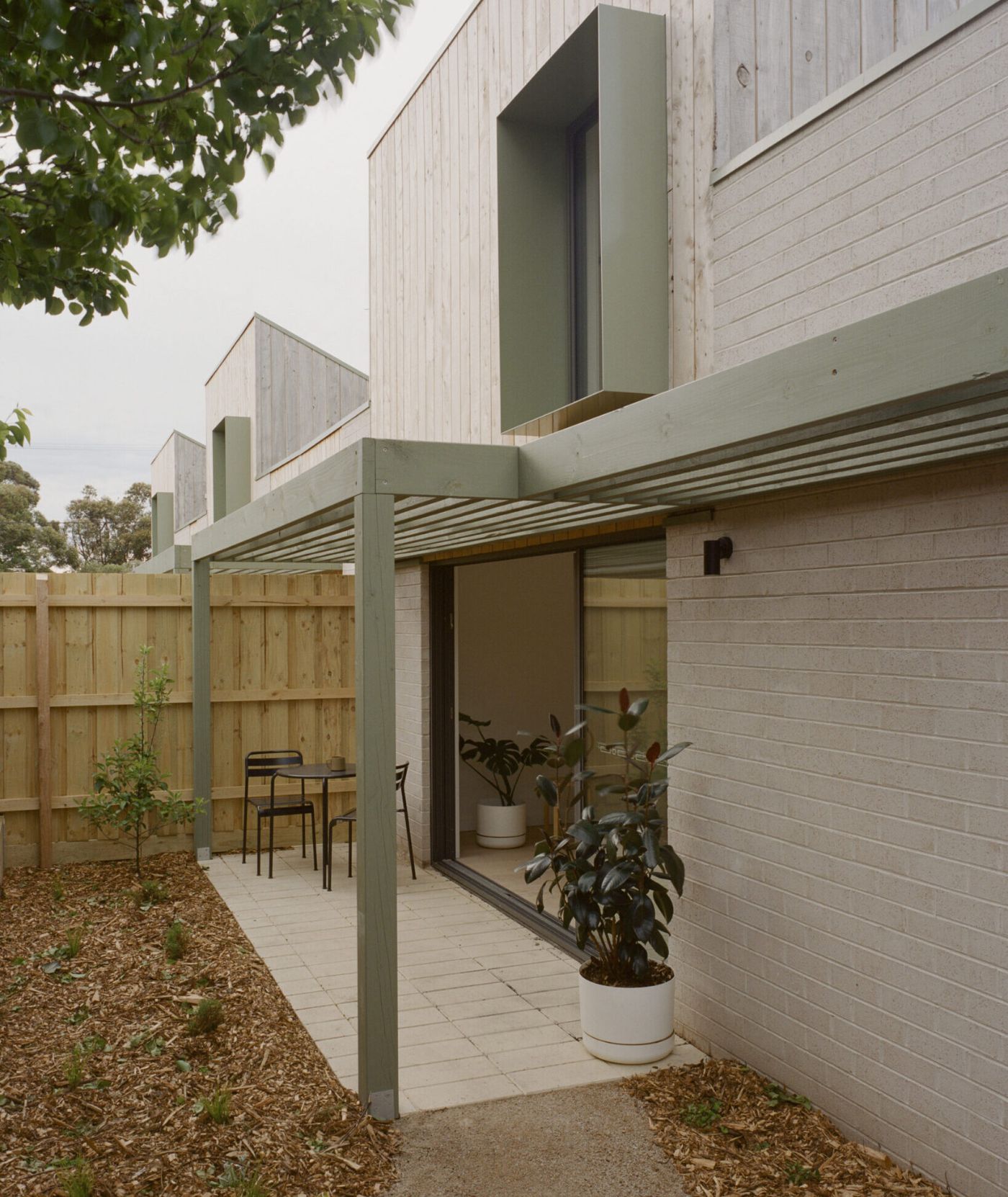 WPI Older Women’s Housing Project by Bright Studio by Studio Bright — A timber-clad dwelling anchors the courtyard threshold, its vertical board cladding ascending to a pitched roof.