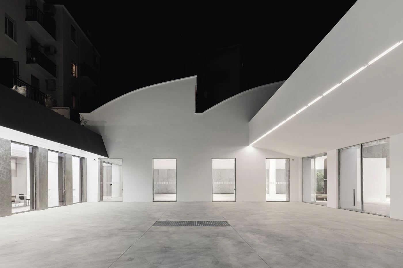 Spazio Maiocchi in Milan — A expansive interior courtyard anchored by sculptural white walls that taper and cantilever overhead, their geometric forms casting sharp shadows across the...