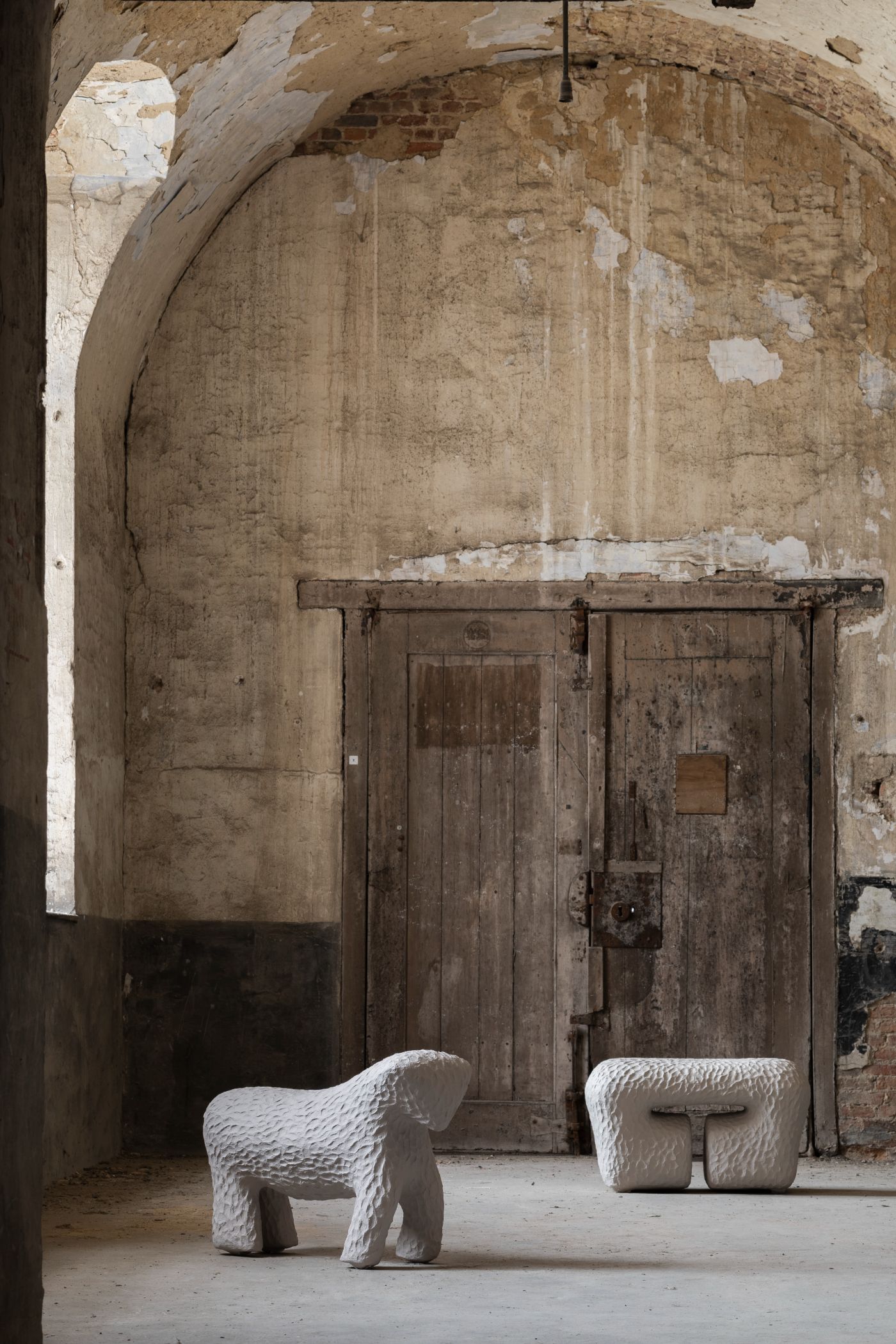 Limited collection by Victoria Yakusha — Two hand-sculpted white stools occupy the floor plane of a vaulted brick interior, their pitted surfaces catching light against weathered masonry walls.