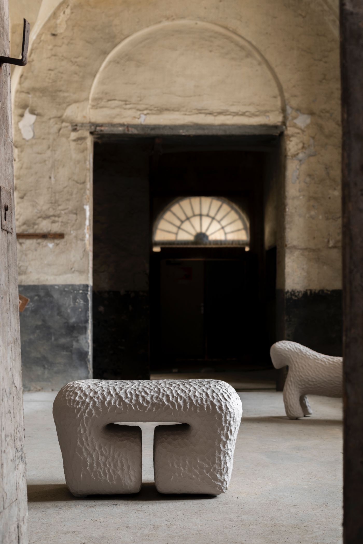 Limited collection by Victoria Yakusha — Two hand-sculpted stone stools occupy the foreground of a vaulted corridor, their surfaces densely textured with incised linear patterns suggesting woven...