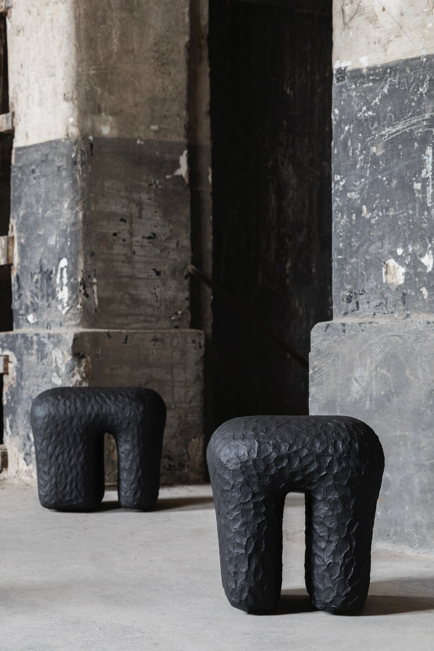 Limited collection by Victoria Yakusha — Two hand-sculpted stools with deeply textured, pitted charcoal surfaces occupy the foreground of an industrial concrete floor.