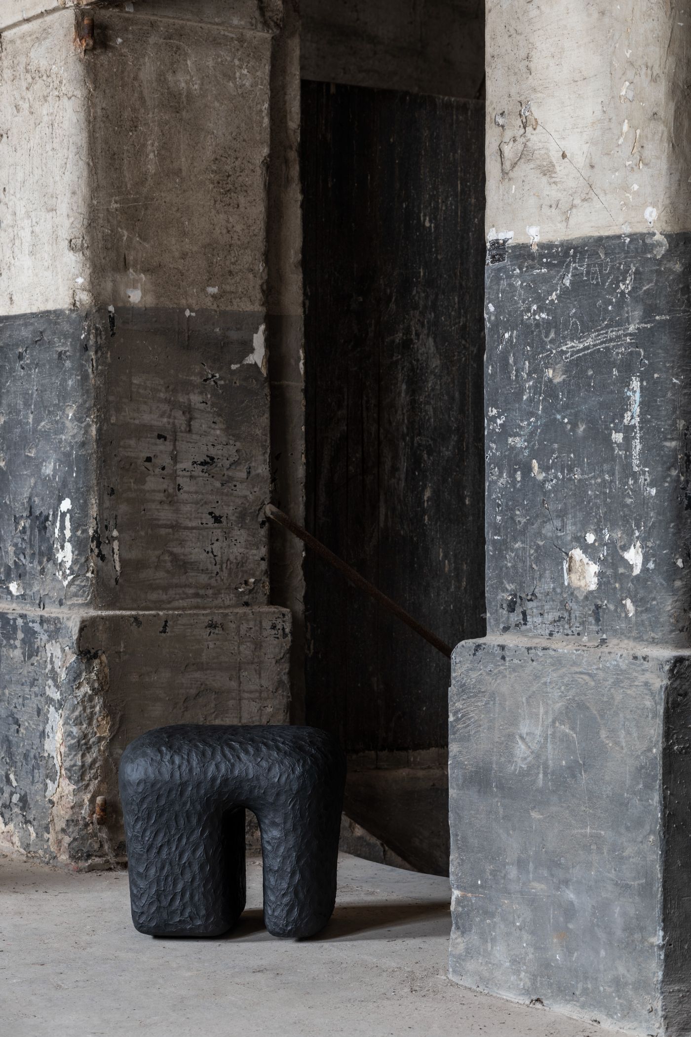 Limited collection by Victoria Yakusha — A hand-sculpted stool in charcoal-black textured material occupies the foreground of a deteriorating industrial interior.