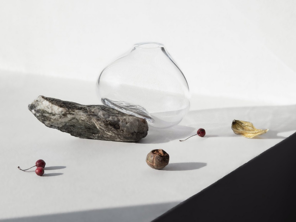 Vase Sculptures by Lætitia Jacquetton — A mouth-blown glass vessel with an irregular, bulbous form rests on a dark weathered stone collected from a riverbed, its surface marked with natural ochre...