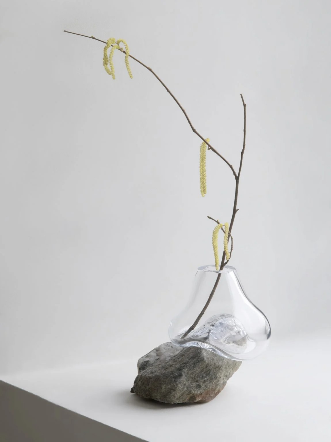 Vase Sculptures by Lætitia Jacquetton — A mouth-blown glass vessel with an irregular, bulbous form sits cradled on a weathered stone base collected from a riverbed, its surface marked by natural...