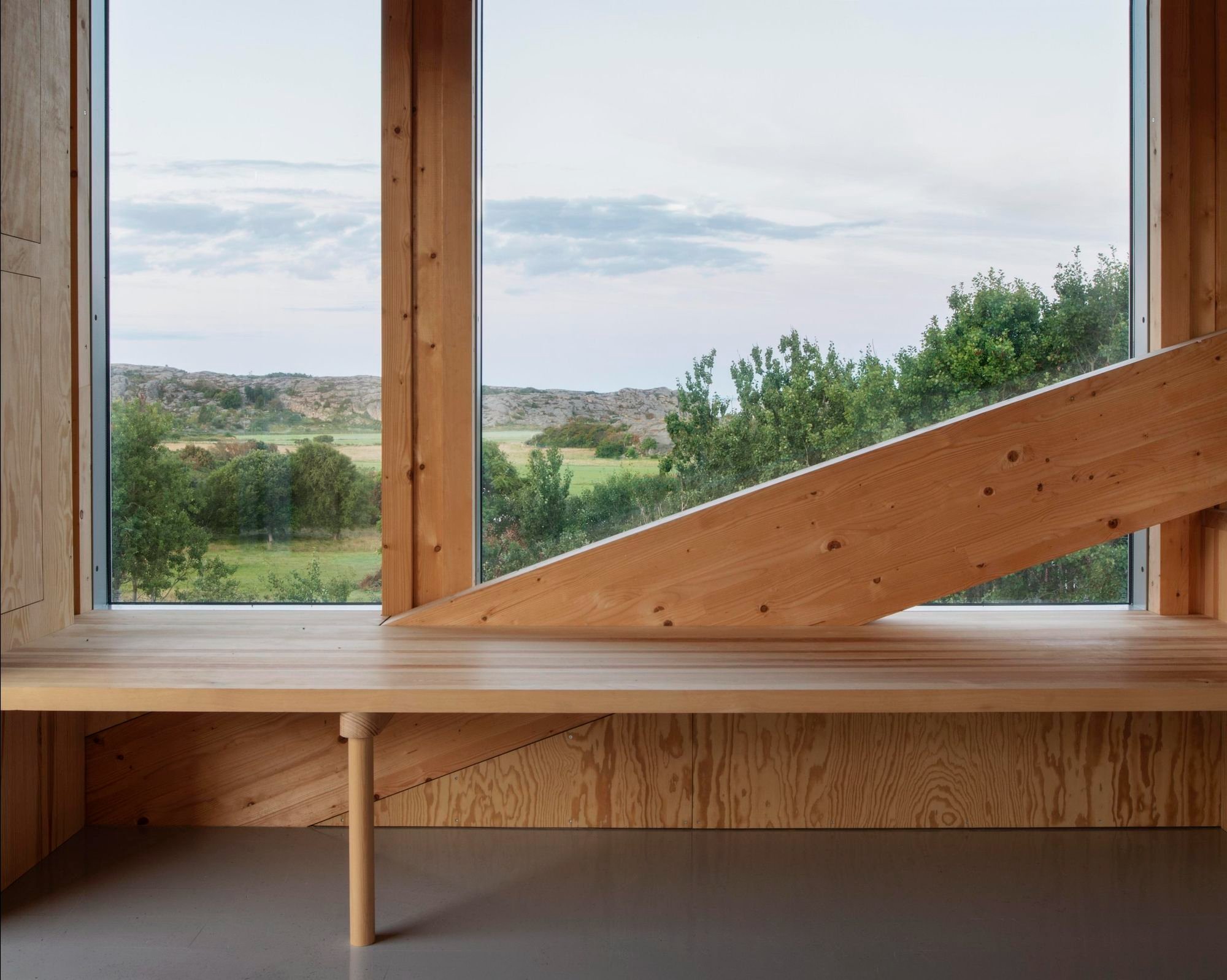 House Hamburgö by Manthey Kula — A timber-framed interior occupies the bridge's threshold, with a long wooden bench and angular bracing beam positioned against expansive glazing.