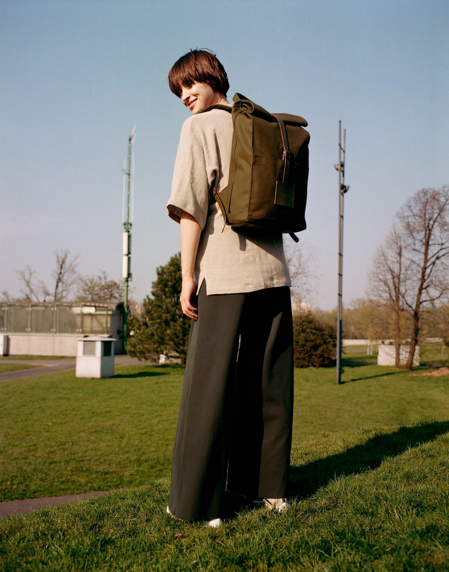 Thisispaper Bags & Backpacks Collection by Thisispaper Studio — A figure stands in profile on manicured grass, wearing a cream-colored V-neck cardigan layered over a beige shirt, paired with black wide-leg trousers and...