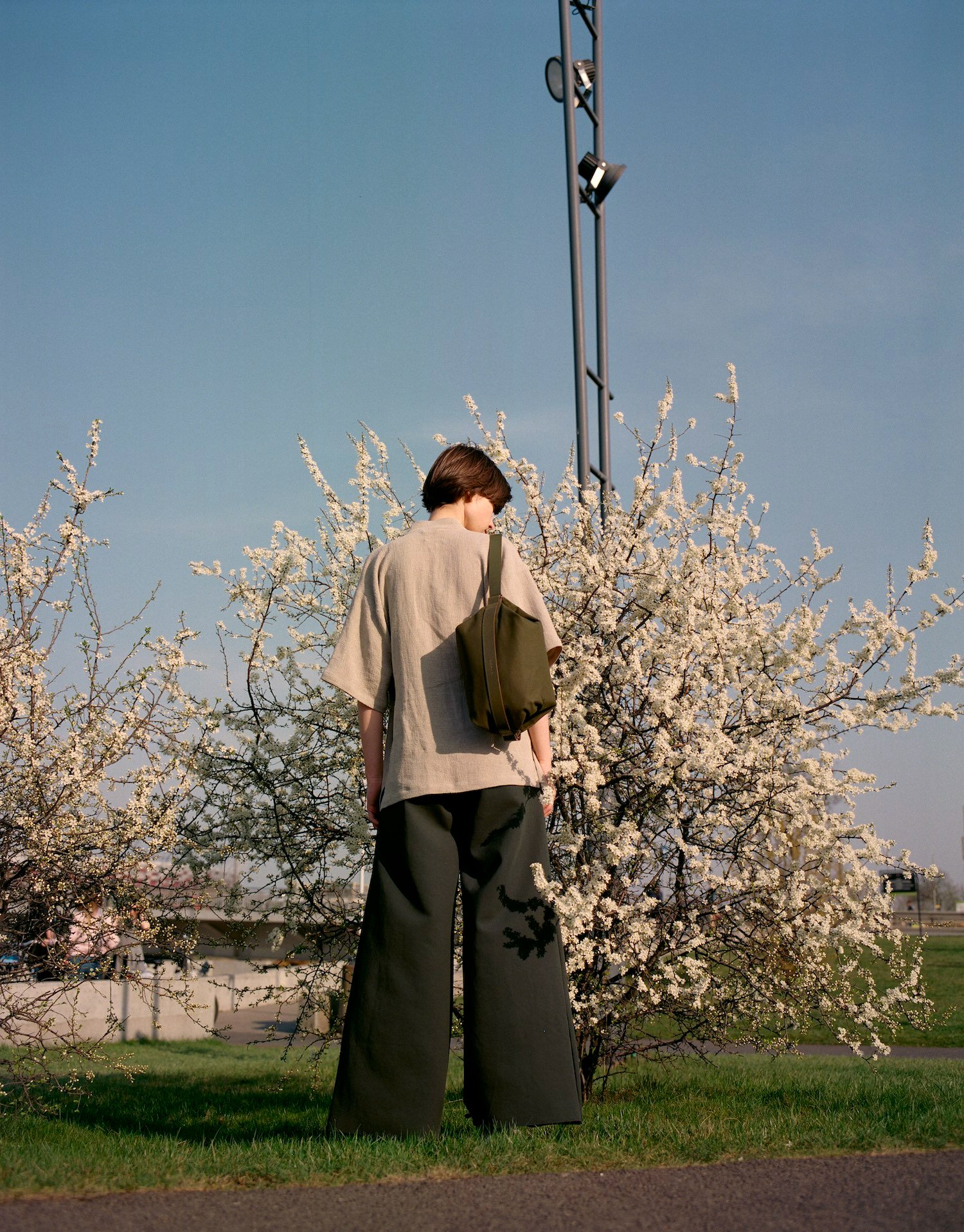 Thisispaper Bags & Backpacks Collection by Thisispaper Studio — A figure stands centered among white-flowering spring branches, wearing a cream-colored knit cardigan and olive canvas utility bag with metal hardware.