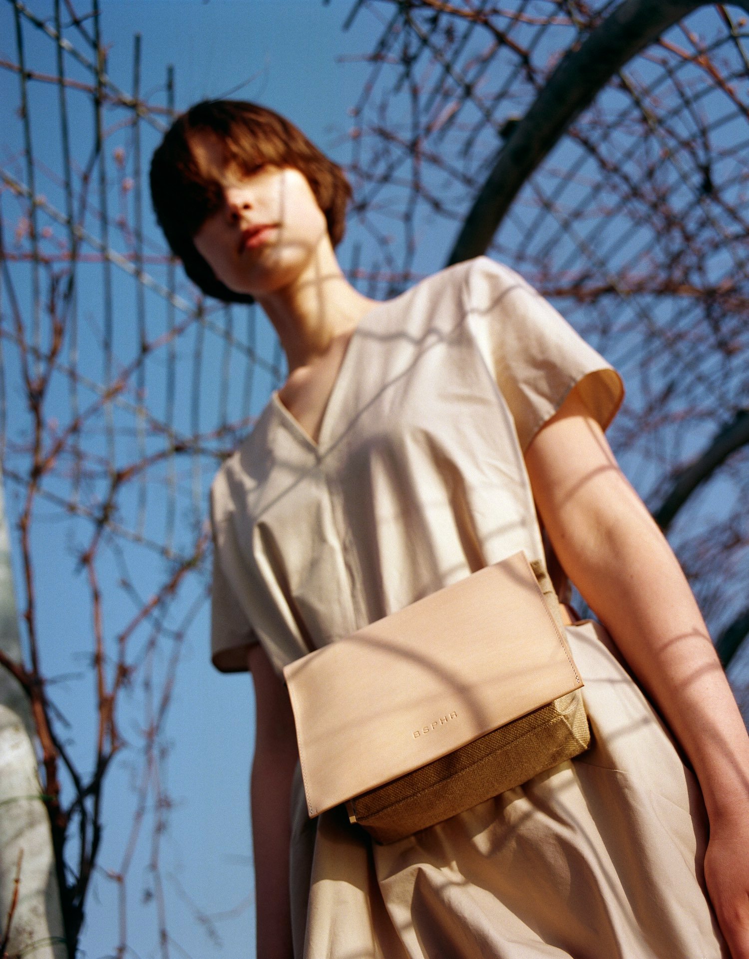Thisispaper Bags & Backpacks Collection by Thisispaper Studio — A figure in cream linen stands beneath spring flowering branches, shot from a low angle against clear blue sky.