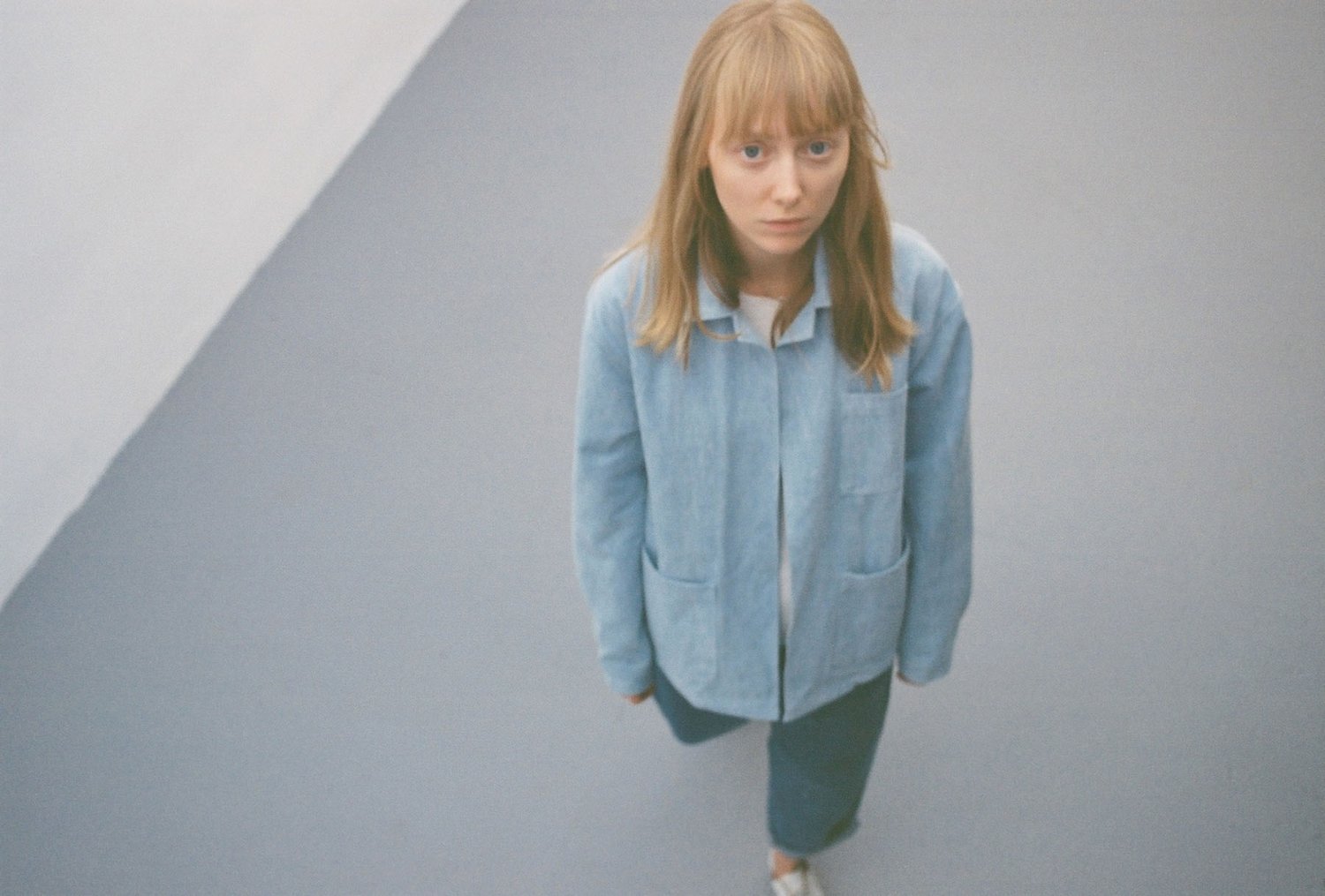 Thisispaper Bags & Backpacks Collection by Thisispaper Studio — A figure stands centered against a neutral white wall in overhead natural light, wearing a light blue chambray shirt jacket with visible chest pockets and a...