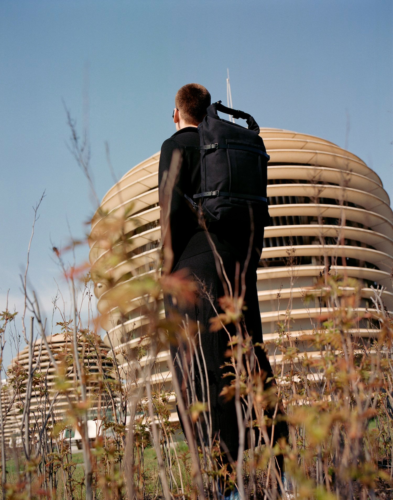 Thisispaper Bags & Backpacks Collection by Thisispaper Studio — A figure wearing a black utility backpack stands amid dry vegetation before a cream-colored residential tower.