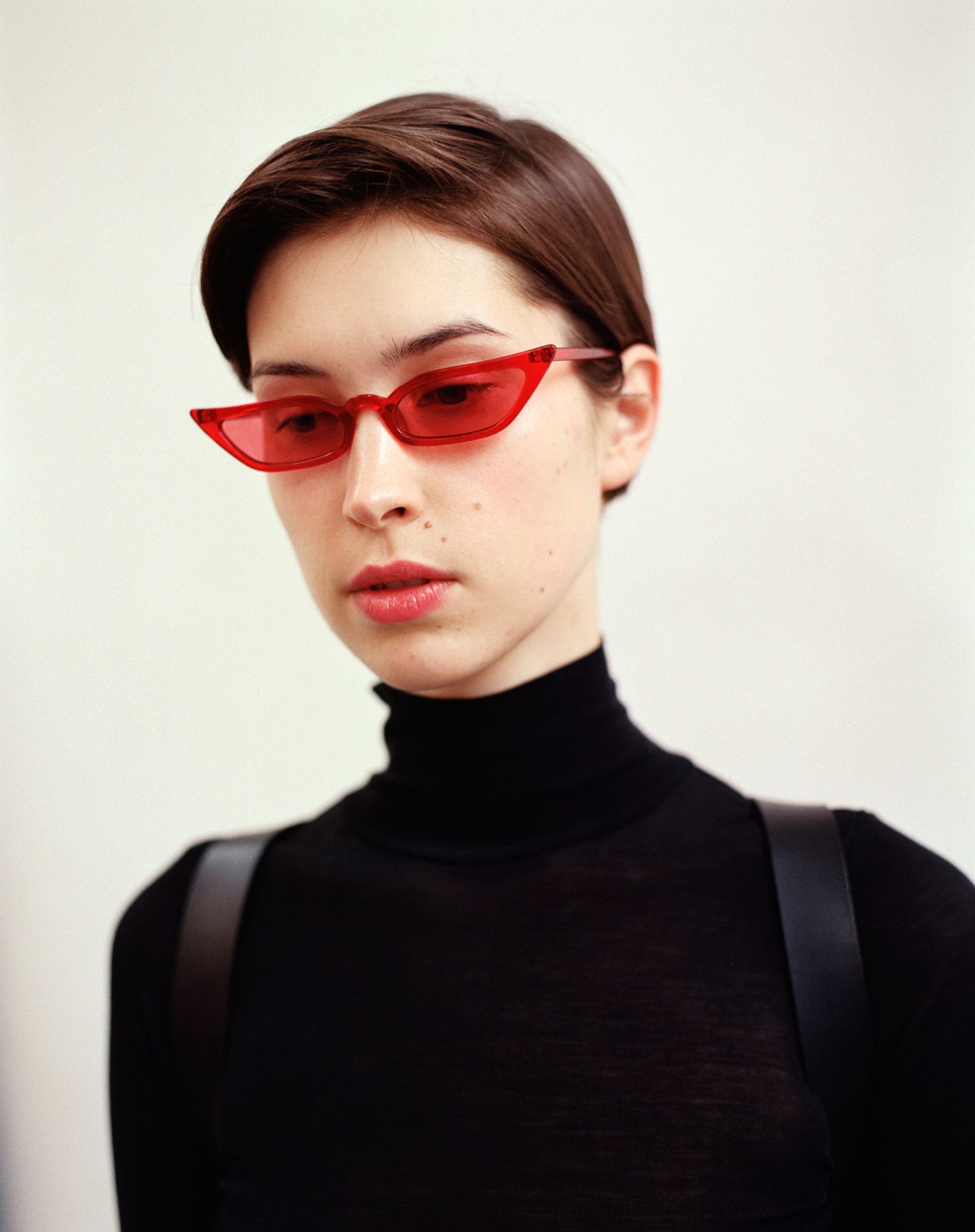 Yenisei by Thisispaper Studio — Portrait photograph featuring a figure in a black turtleneck wearing red tinted cat-eye sunglasses against a neutral cream background.