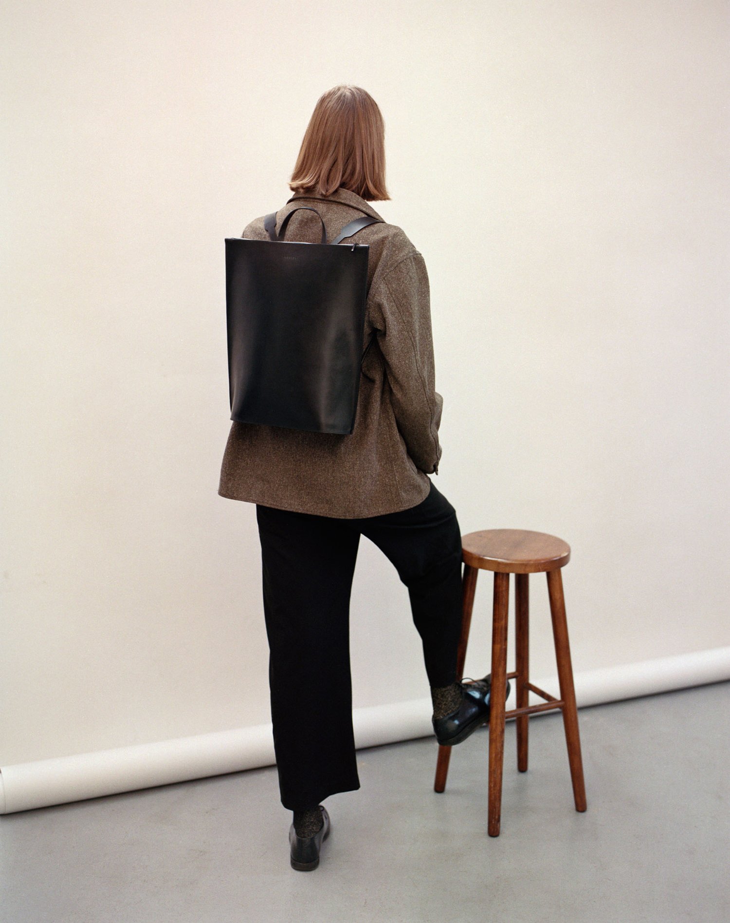 Yenisei by Thisispaper Studio — A figure stands in three-quarter view wearing a textured grey-brown wool jacket with visible weave, black trousers, and black platform shoes.