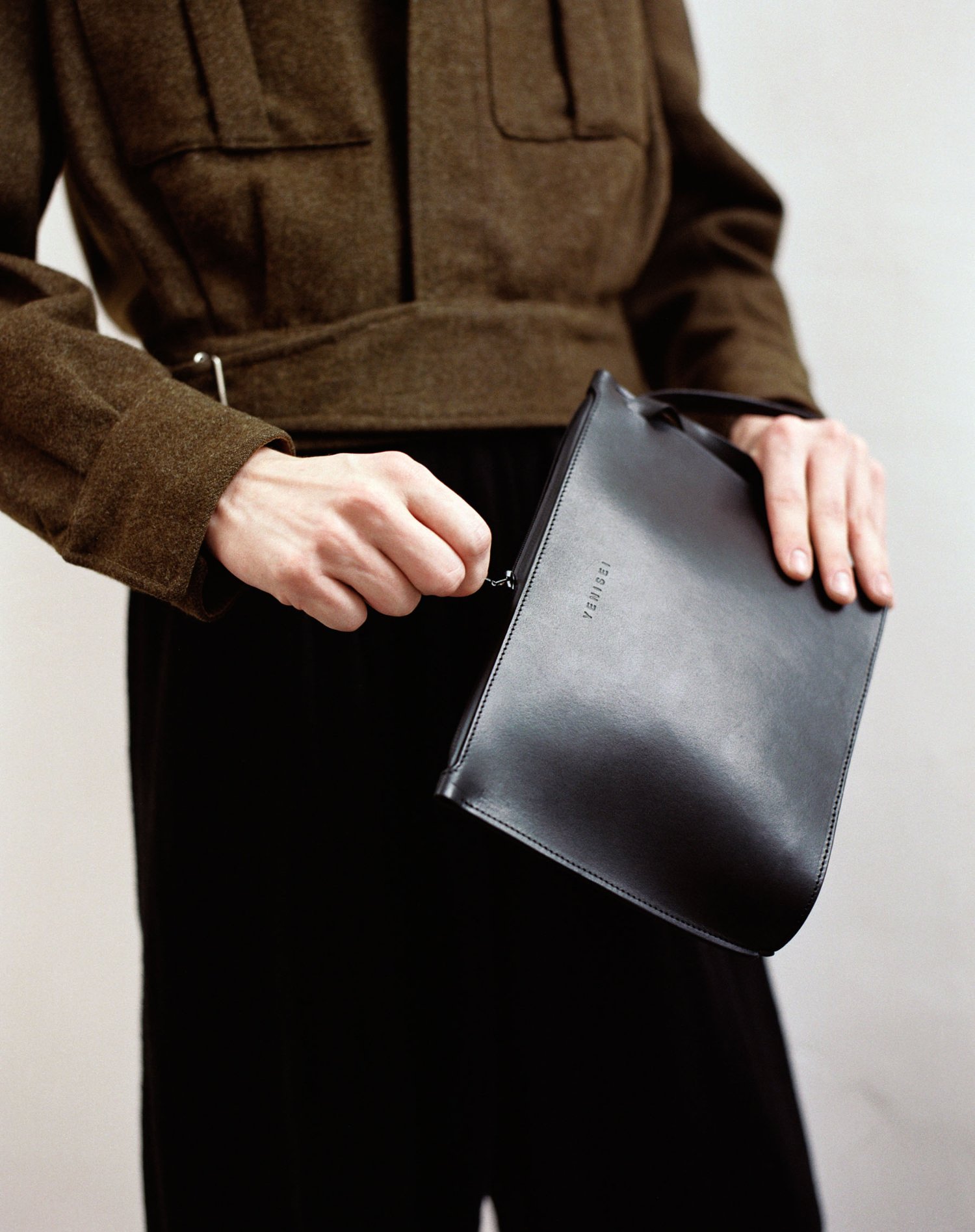 Yenisei by Thisispaper Studio — A product photograph isolating a black leather clutch held by hands emerging from a brown wool jacket.