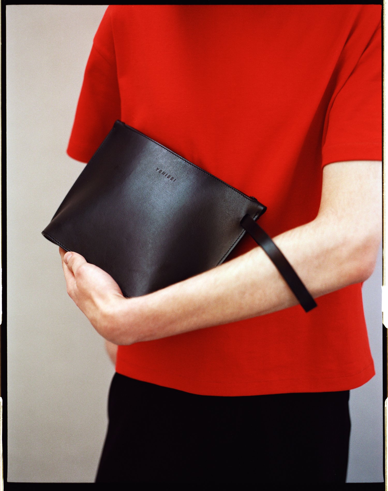 Yenisei by Thisispaper Studio — A black leather clutch with minimal branding detail is held against the torso of a figure wearing a vibrant red short-sleeved top and black trousers.