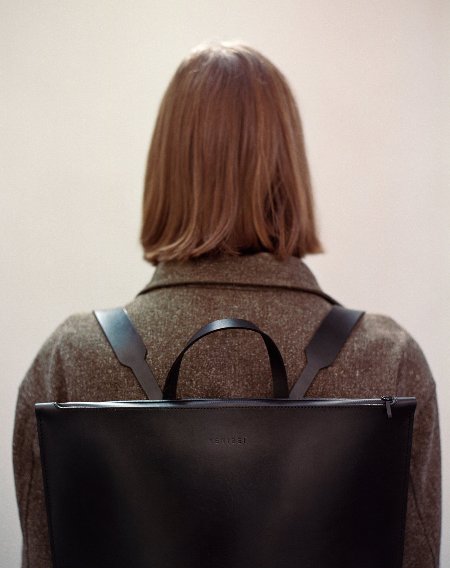Yenisei by Thisispaper Studio — Rear view of a figure wearing a textured grey-brown wool jacket with visible weave, carrying a black leather tote backpack with dual straps and minimal...