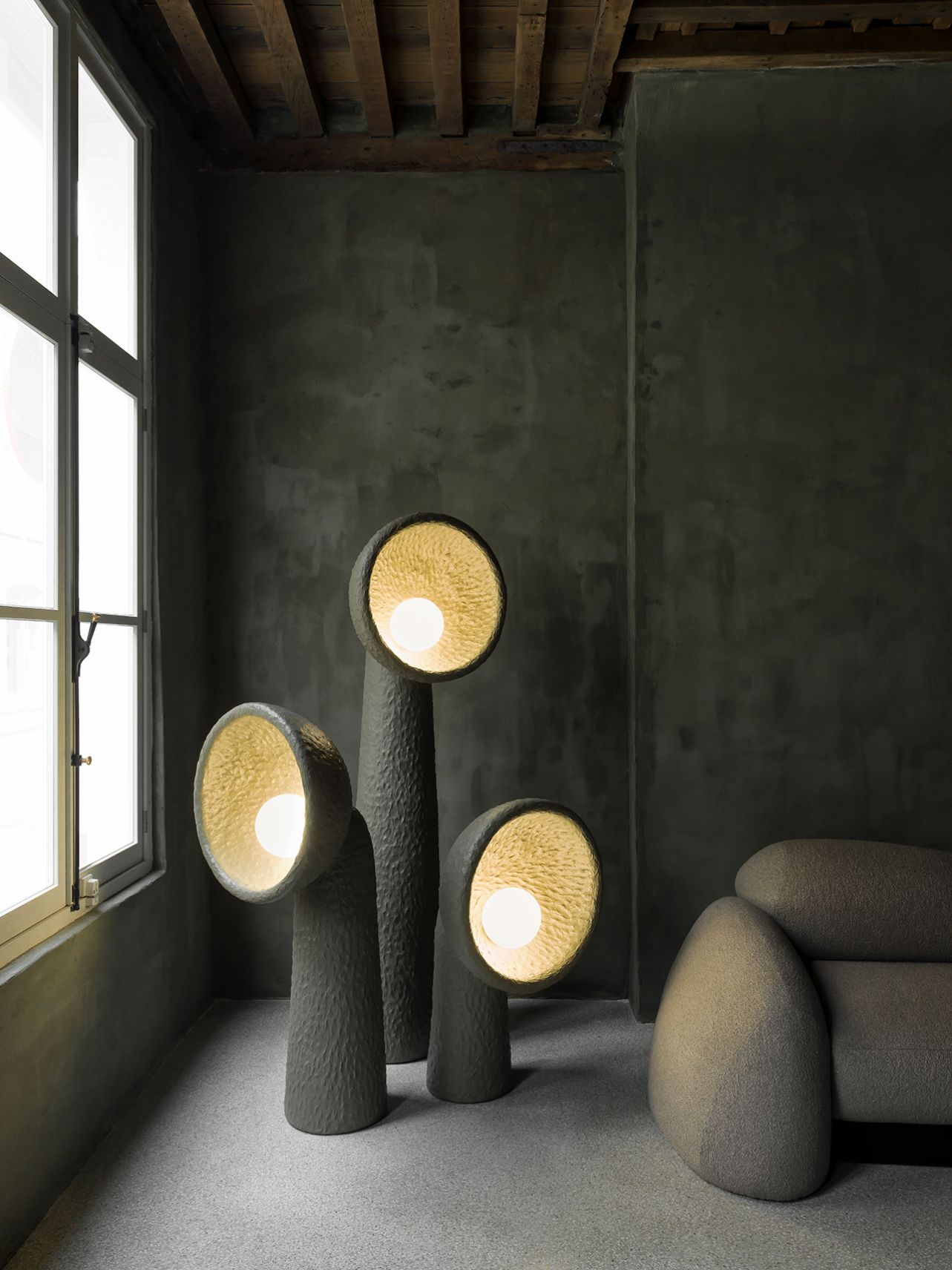 Faina Gallery by Yakusha Studio — Three monolithic ceramic vessels with textured surfaces occupy the gallery floor, their cylindrical forms topped with circular openings housing warm golden...