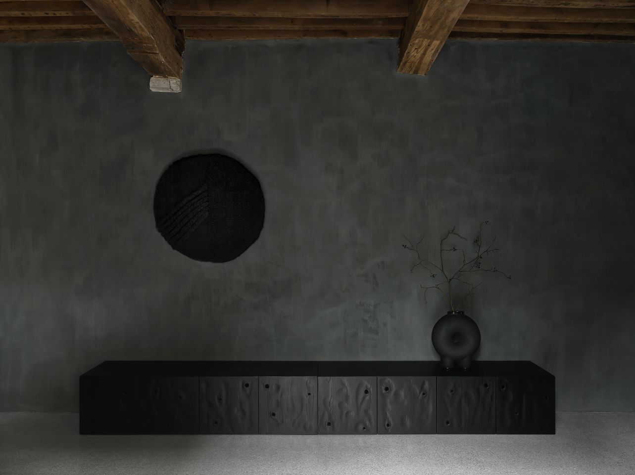Faina Gallery by Yakusha Studio — A minimalist gallery interior anchors a black timber credenza against a hand-rendered charcoal-grey wall with visible trowel marks and surface irregularity.