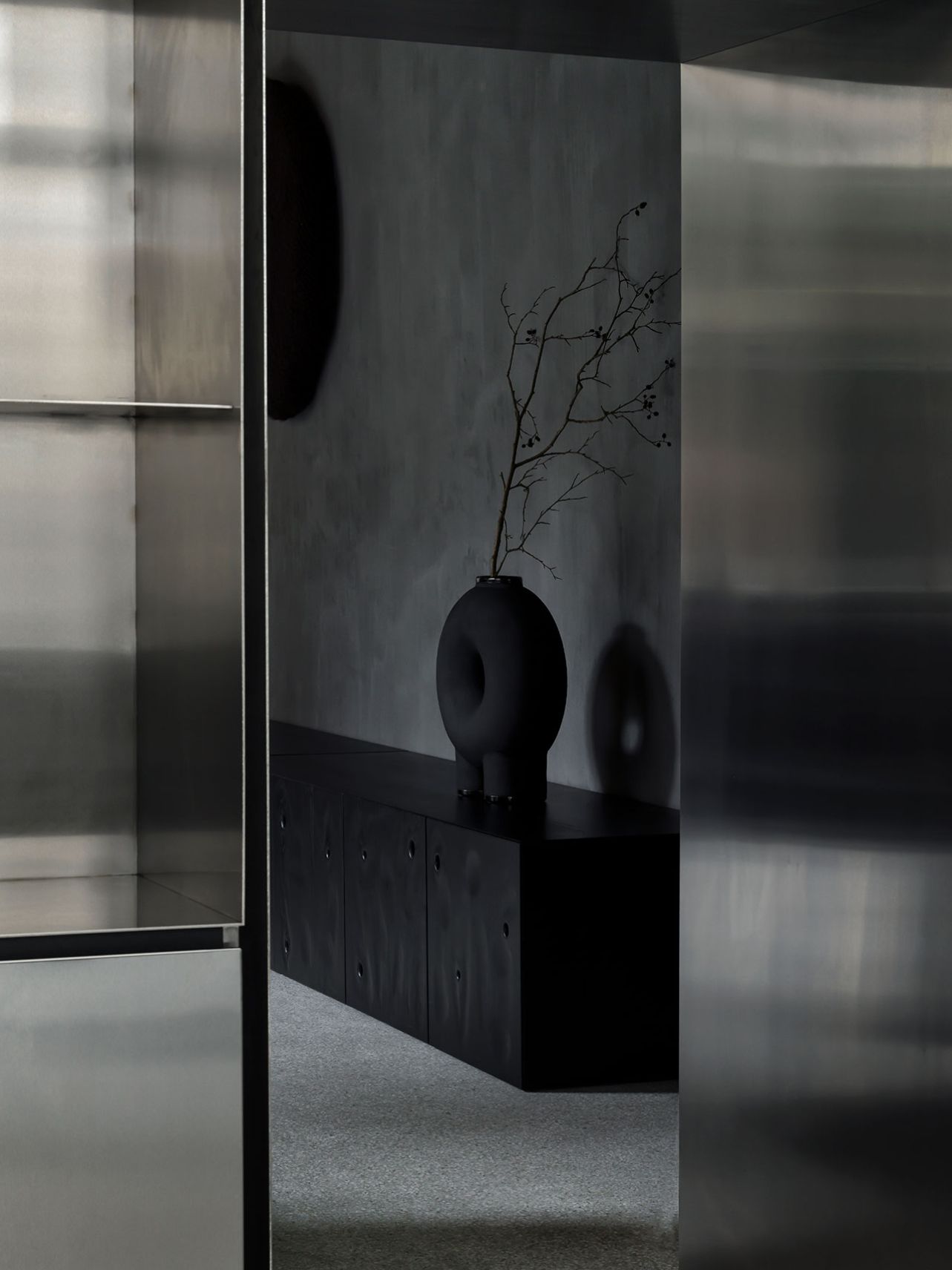 Faina Gallery by Yakusha Studio — View through a gridded glass door frame into the gallery interior, where a black ceramic vessel with organic, bulbous form sits atop a dark timber plinth.