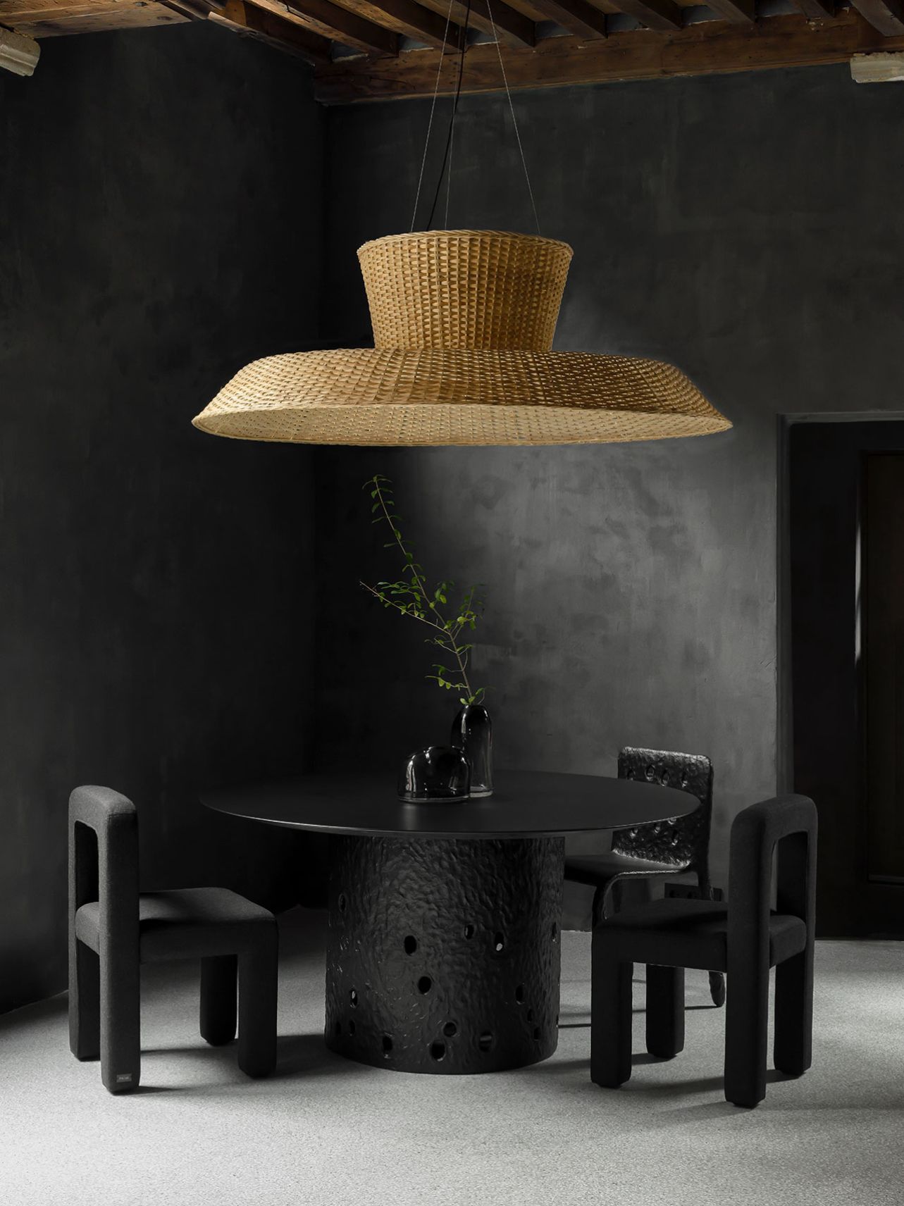 Faina Gallery by Yakusha Studio — A dining ensemble anchors the gallery's austere interior: a cylindrical black ceramic table base with organic surface perforations sits beneath a woven...
