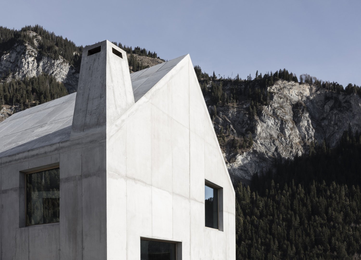 The Trin Cabin By Schneller Caminada Architects — A three-story concrete dwelling set against forested Alpine slopes, its monolithic white facade punctuated by three asymmetrically positioned window openings.