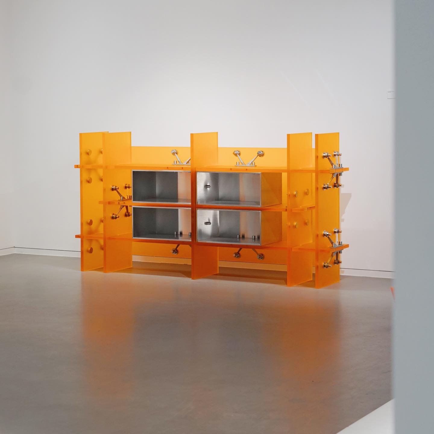 Mixed Public-Private Boundary collection by Cometabolism Studio — A volumetric shelving unit constructed from repurposed industrial components, finished in high-saturation orange with visible mechanical hardware across its...