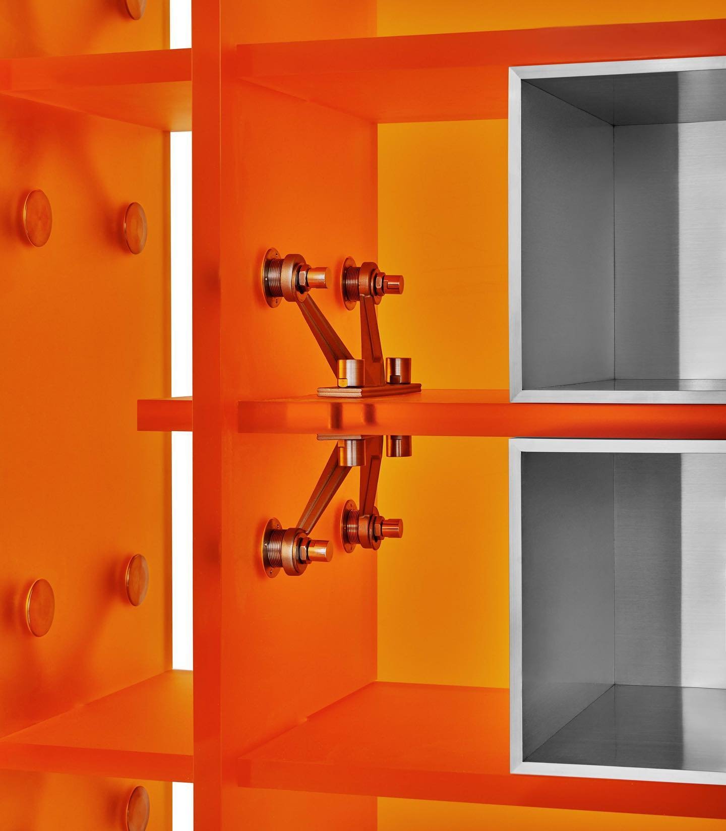 Mixed Public-Private Boundary collection by Cometabolism Studio — Two welded steel mechanical assemblies mounted on a vivid orange-rendered wall, their brass and copper components in sharp relief.