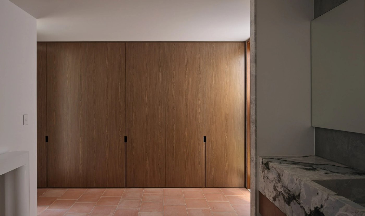 Chelmer River House by Furminger by Christopher Furminger — A timber-clad interior wall system dominates the frame, composed of three vertical panel sections with recessed black handles creating a linear rhythm...