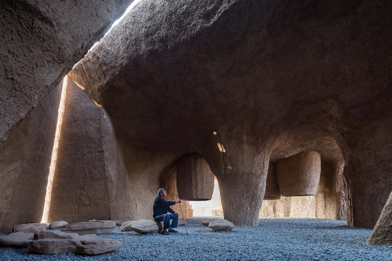 Holy Fire Lit Place in China by URBANUS — A solitary figure in dark clothing crouches within the cavernous interior of the Xihoudu museum, dwarfed by massive earthen walls carved from monolithic...