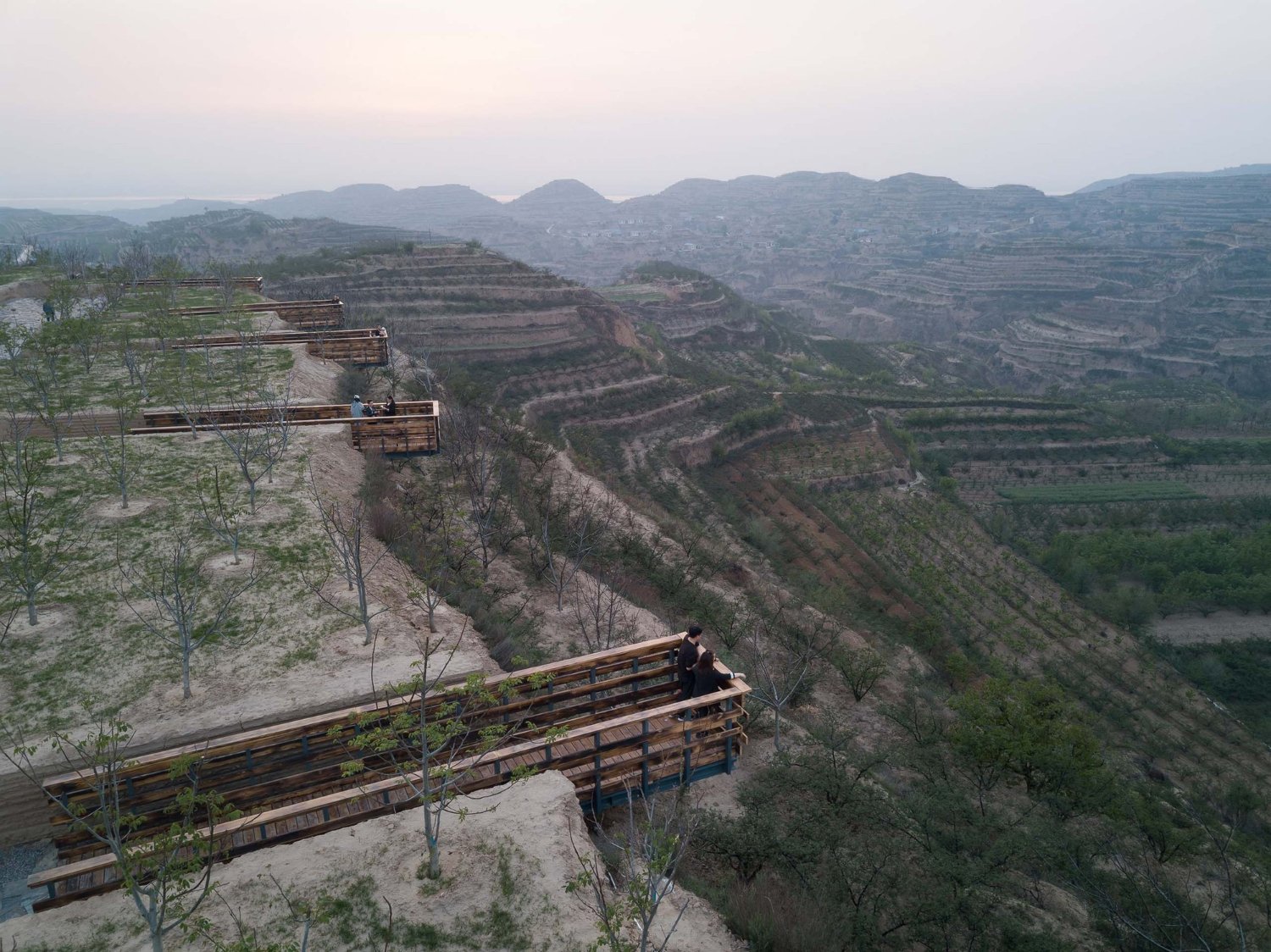 Holy Fire Lit Place in China by URBANUS — Timber-framed viewing platforms cantilever dramatically from the earthen cliff face, their rust-patinated steel structural elements anchoring into the...