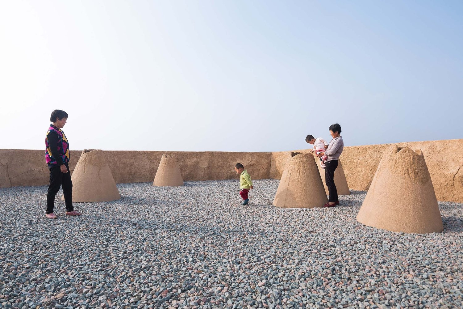 Holy Fire Lit Place in China by URBANUS — Children and adults occupy a gravelly courtyard scattered with monolithic earthen cone structures.