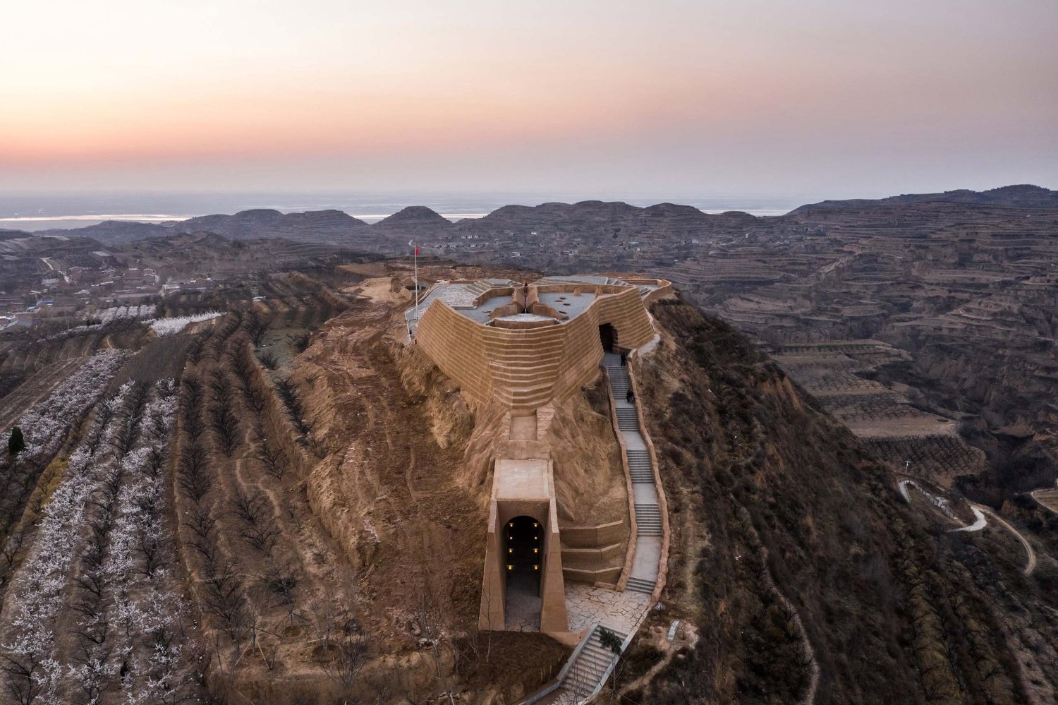 Holy Fire Lit Place in China by URBANUS — Aerial view of the Xihoudu museum captured at dawn, revealing the monolithic earthen structure carved into a ridge overlooking the Yellow River valley.