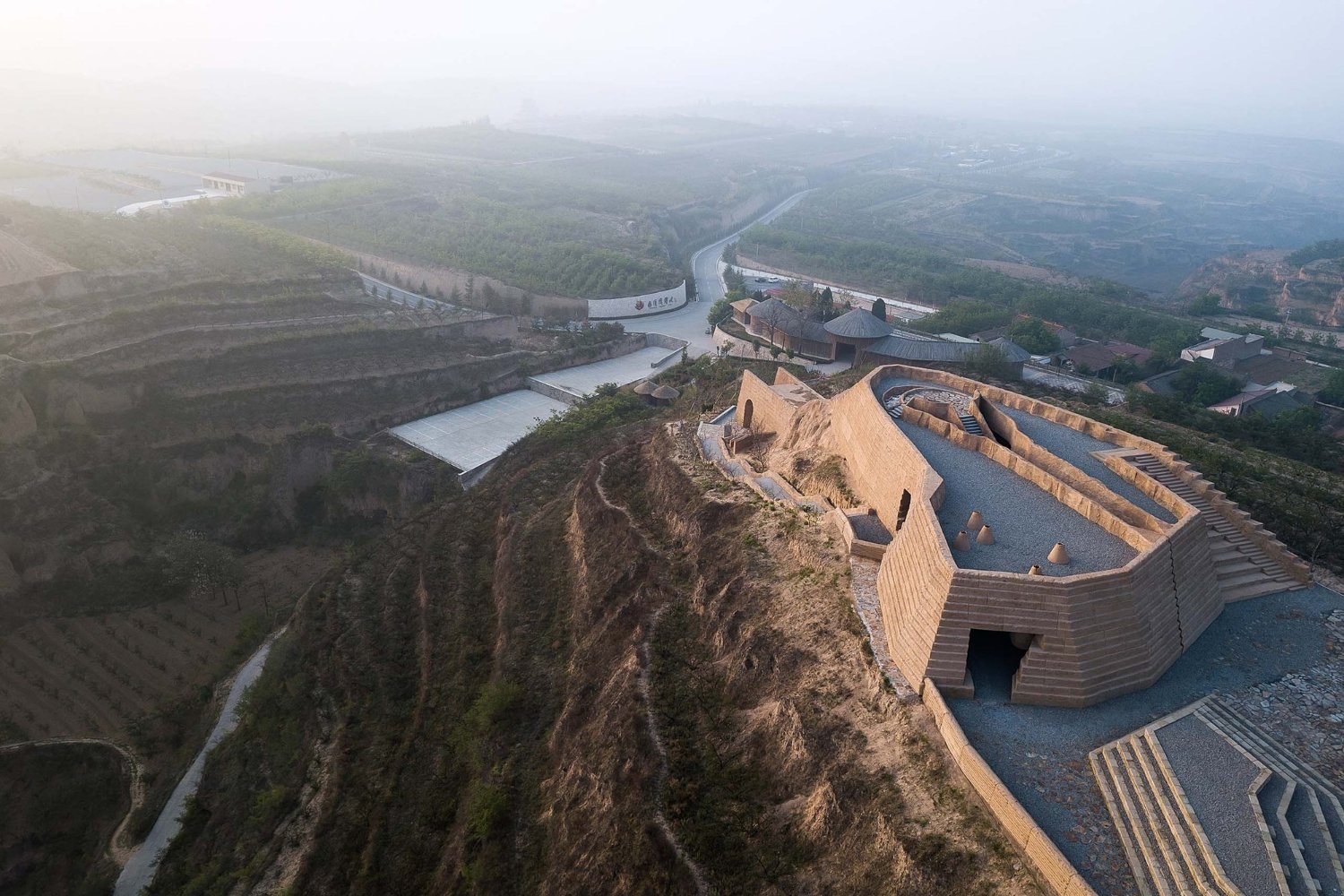 Holy Fire Lit Place in China by URBANUS — Aerial view of the monolithic earthen structure carved into the cliff edge, revealing the tree-form massing with radiating branch-like apertures that...