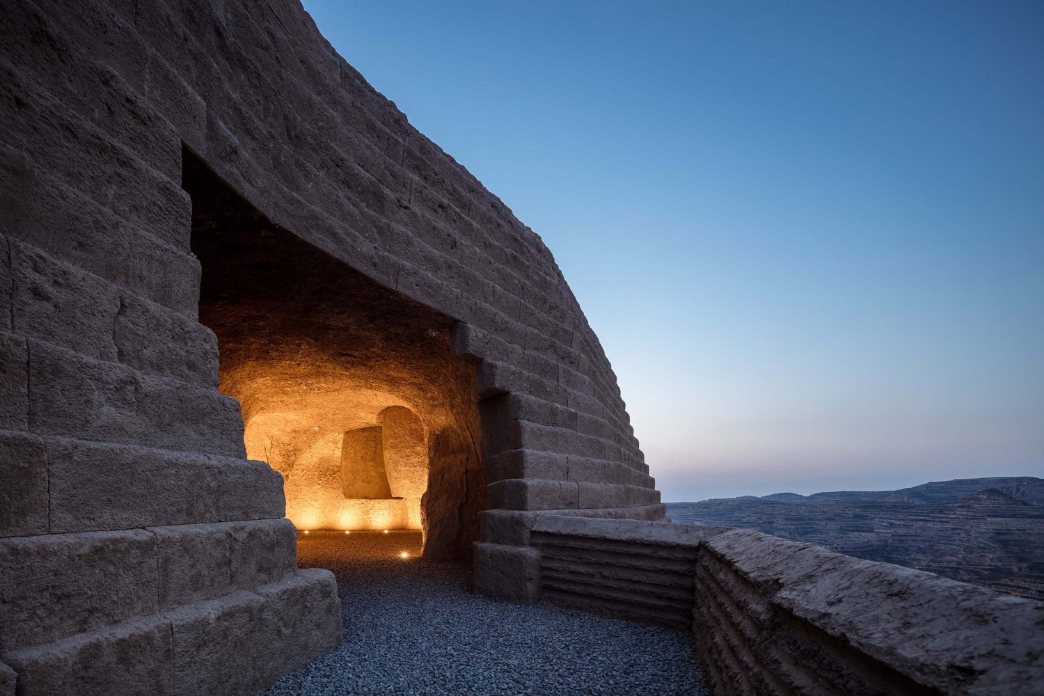 Holy Fire Lit Place in China by URBANUS — A monolithic earthen chamber carved from rammed-earth mass, illuminated from within by warm artificial light.
