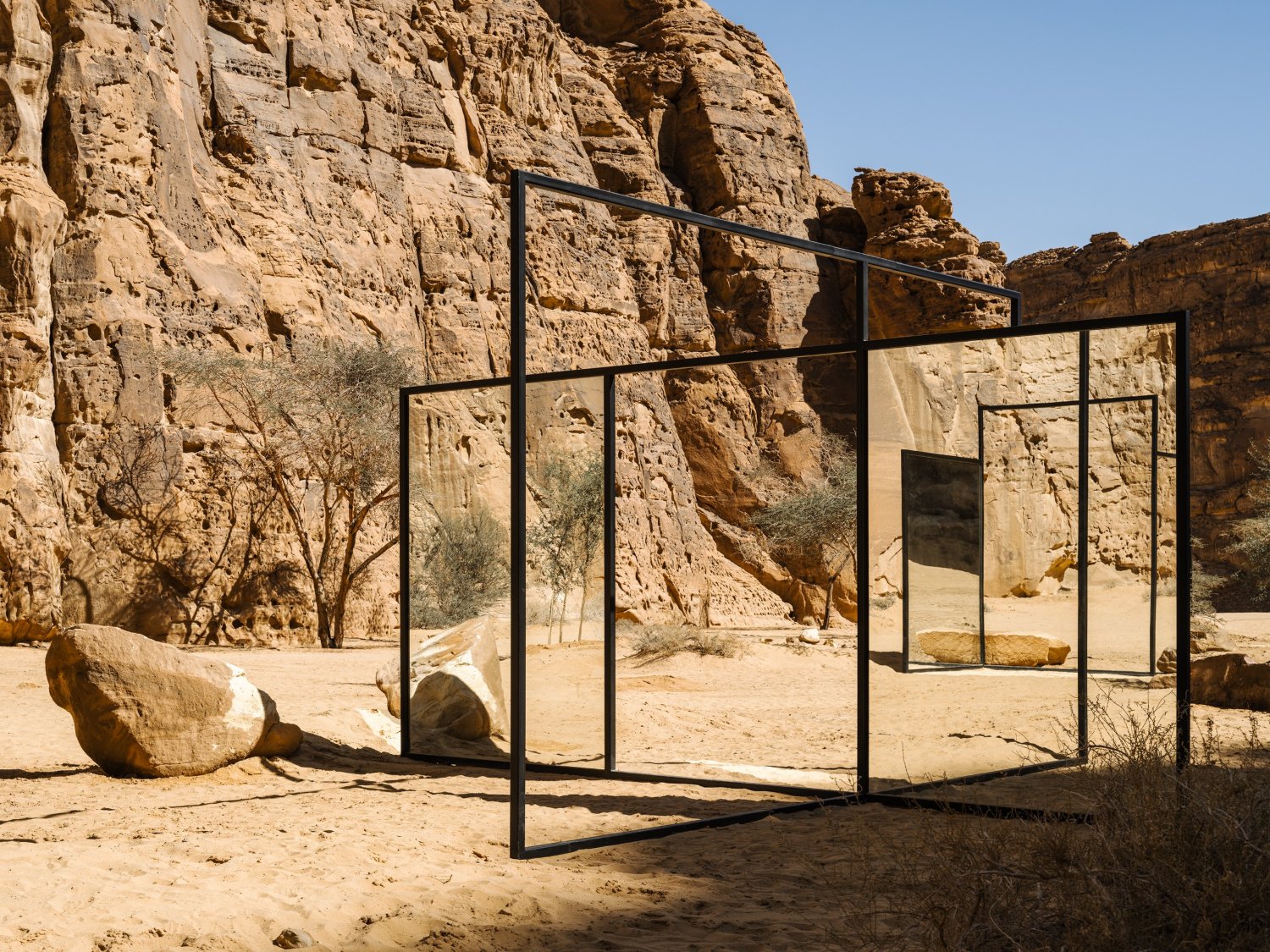 Desert X AlUla 2022 Group Exhibition — A monumental steel-frame installation occupies the desert floor, its minimalist geometry sharply contrasting the weathered sandstone cliff face behind.